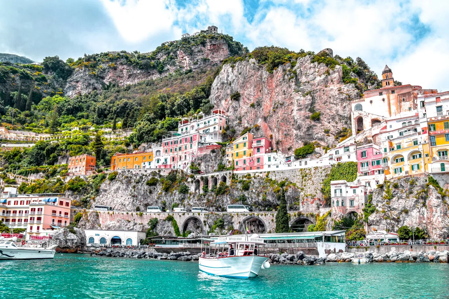 Amalfi Kıyısı'nın dik yamaçlarına oyulmuş, rengarenk evlerin sıralandığı ve kıyı boyunca uzanan kemerli yolların göründüğü bu eşsiz manzarada, turkuaz denizde bir tekne ilerliyor ve bölgeye nasıl gidilebileceğine dair büyüleyici bir görsel sunuyor.