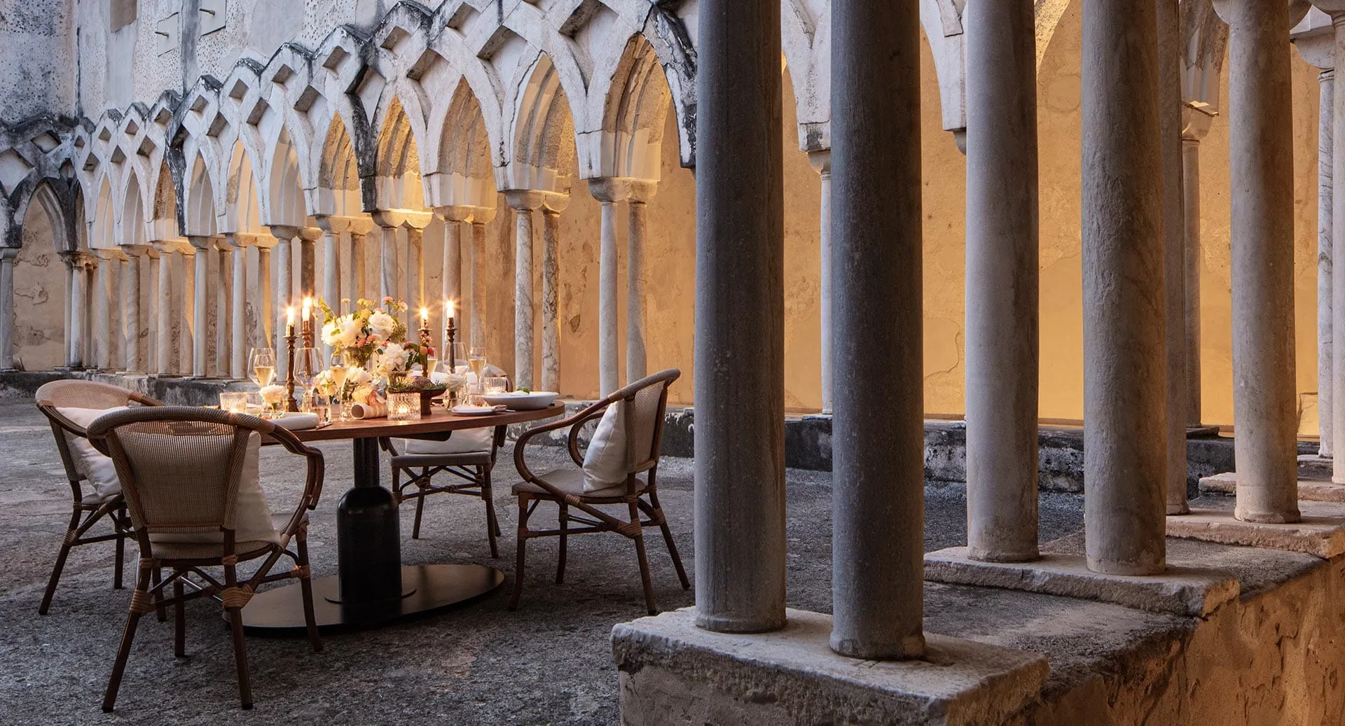 Anantara Convento di Amalfi Grand Otel'in tarihi taş revaklı avlusunda, zarif sandalyelerle çevrili, mum ışıkları ve taze çiçeklerle özenle süslenmiş yuvarlak bir yemek masası, otelin lüks ve büyüleyici Akdeniz atmosferini yansıtmaktadır.