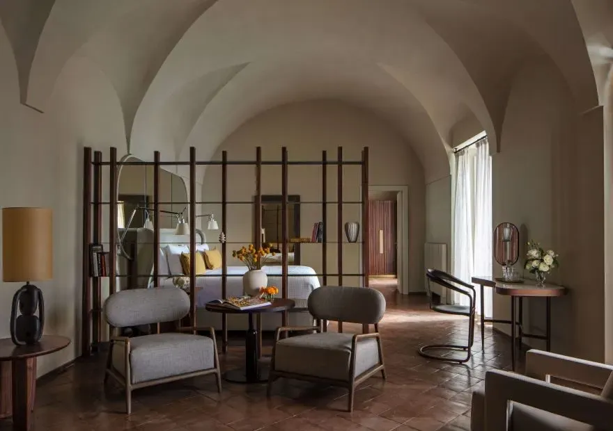 Anantara Convento di Amalfi otelindeki tonozlu tavanlı, terasit fayans zeminli geniş ve lüks bir odada, ahşap bir paravanla ayrılmış yatak alanının önünde modern koltuklar ve çiçeklerle süslenmiş masalar içeren zarif bir oturma alanı sakin bir atmosfer sunuyor.