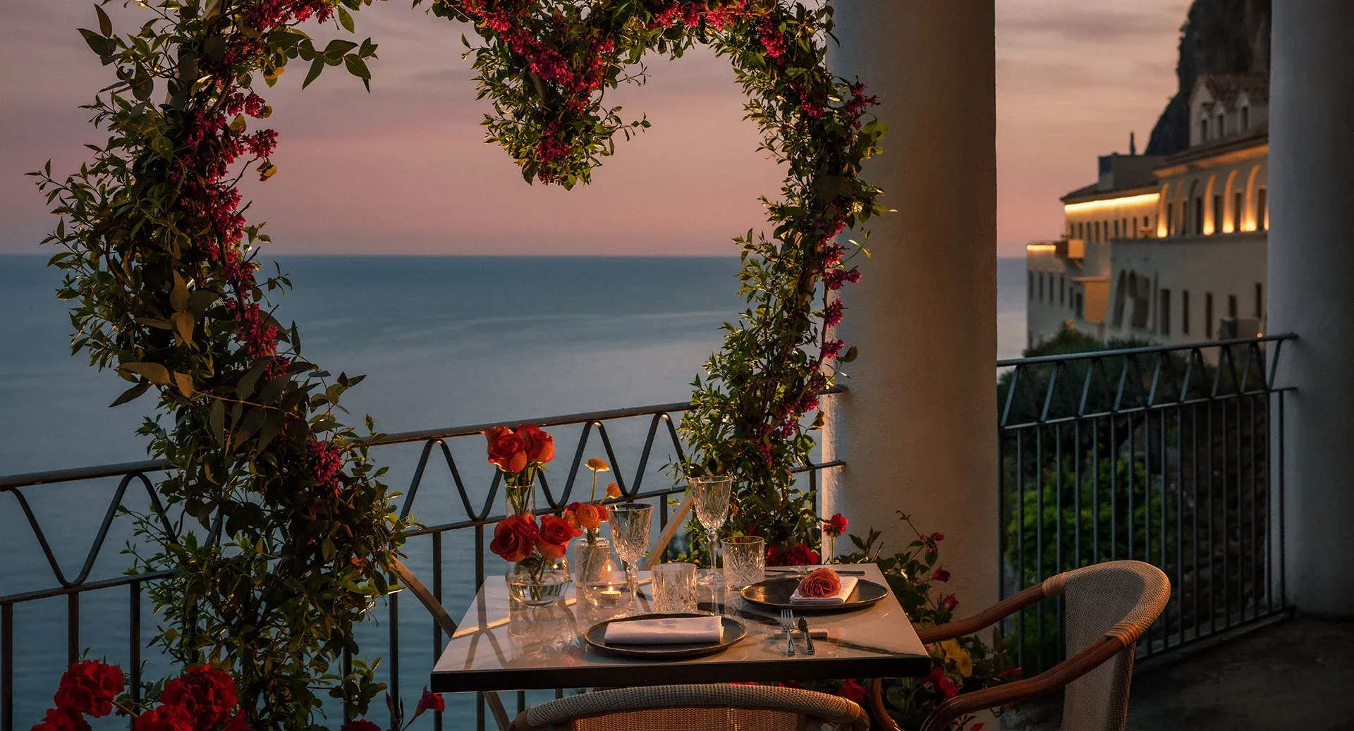 Gün batımında, denize nazır lüks bir balkonun romantik atmosferinde, kalpli çiçeklerle süslenmiş demir korkulukların ardında, mum ışığıyla aydınlatılmış, iki kişilik özel bir akşam yemeği masası; zarif porselenler, kristal kadehler ve vazoda canlı turuncu güllerle donatılırken, uzakta ışıkları yanan sahil binaları ve pastel tonlardaki gökyüzü büyüleyici bir manzara sunuyor.