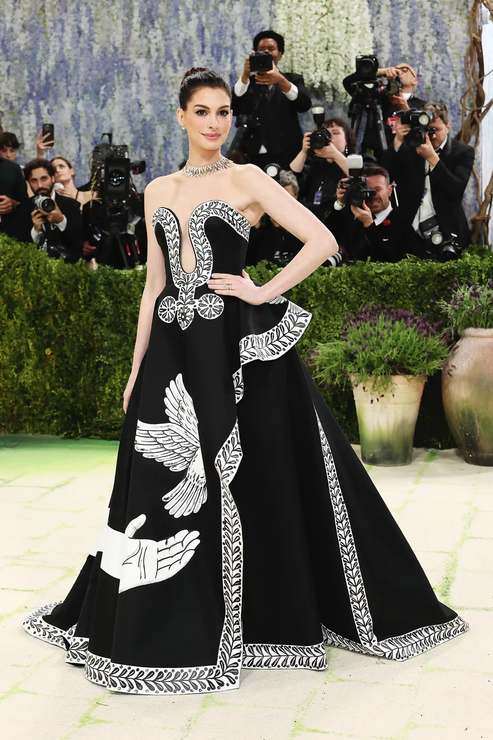 Anne Hathaway, 2026 Met Gala Kırmızı Halısı'nda, straplez siyah elbisesinin eteklerinde beyaz güvercinler ve ellerin sembolik motifleri ile sanatsal yaprak desenlerinin dikkat çektiği göz alıcı bir kostümle, arkasında fotoğrafçı kalabalığı varken zarifçe poz veriyor.