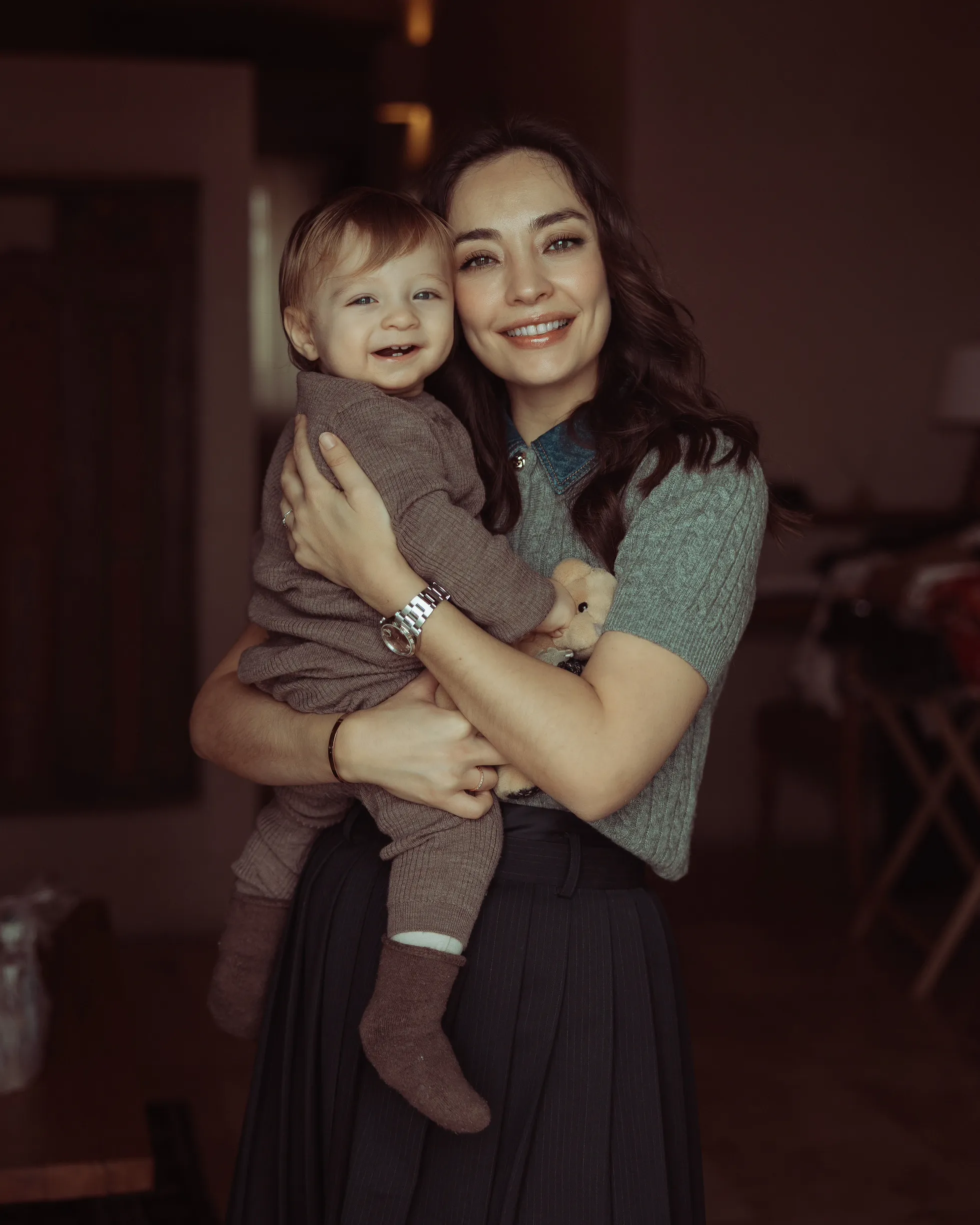 Oyuncu Zeynep Tuğçe Bayat, gri örgü kazak ve koyu renk pilili etekle, neşeyle gülümseyen açık renk saçlı oğlu Atlas'ı kucağında şefkatle tutarken kameraya bakıyor ve anneliğin ona sunduğu yeni denge ve güven duygusunu içtenlikle yansıtıyor.
