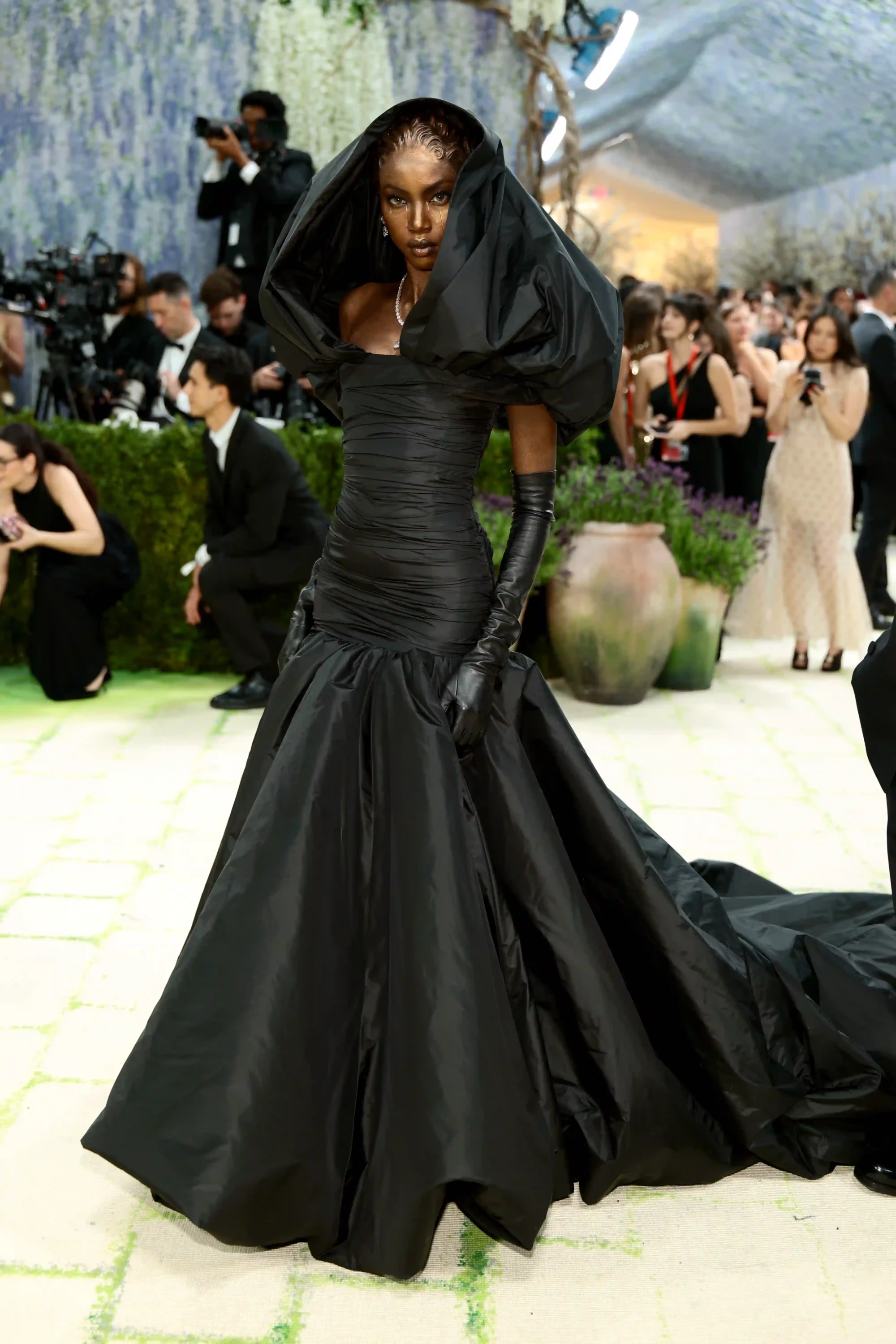 Anok Yai'nin 2026 Met Gala kırmızı halısında giydiği, başını saran kapüşonlu, kabarık kollu, büzgülü bedeni ve yere kadar uzanan hacimli eteğiyle heykelsi bir görünüm sunan tamamen siyah kostümü, altın rengi makyajı ve pırlanta takılarıyla dikkat çekiyor.