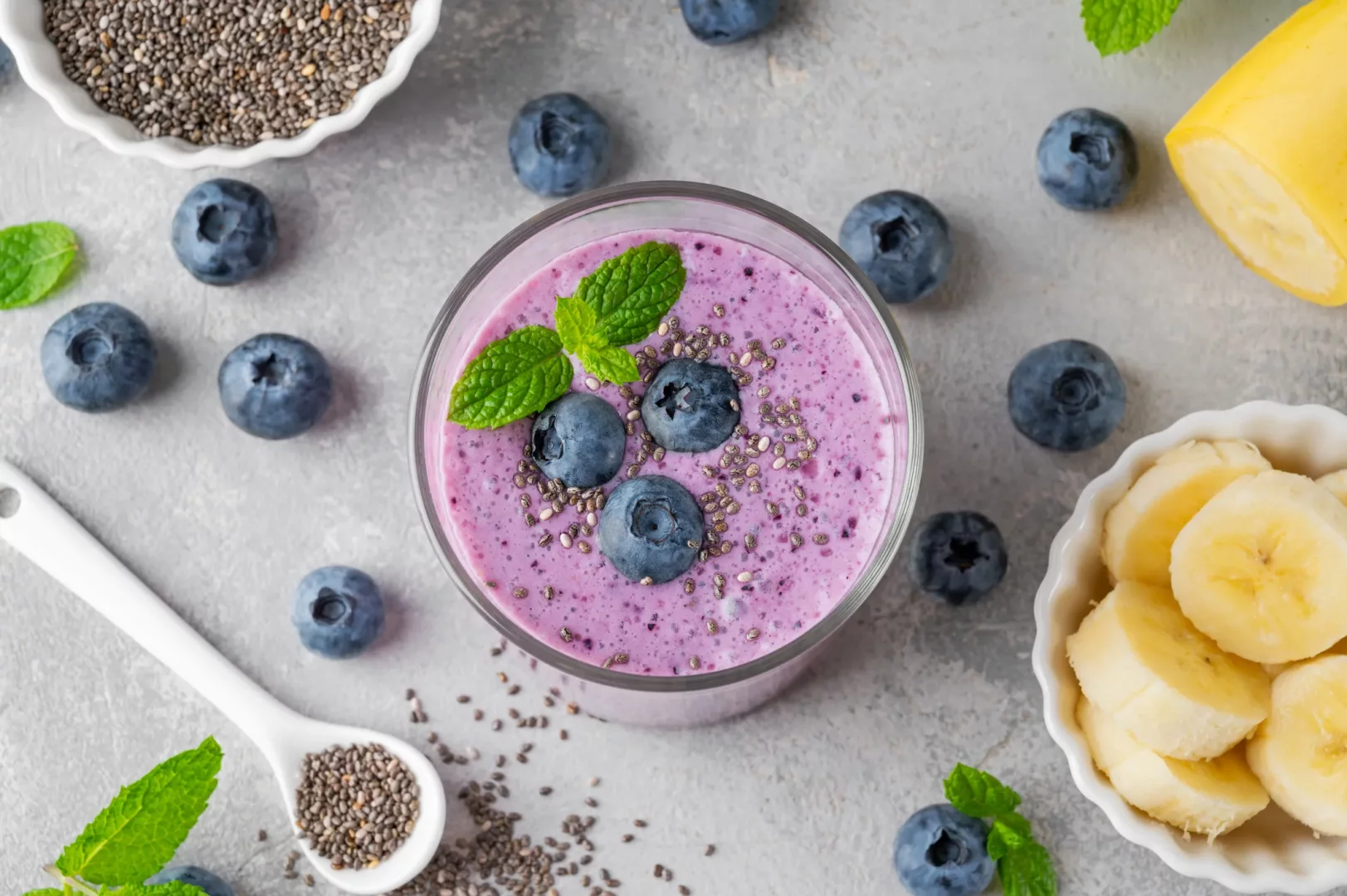 Gri bir zeminde üstten çekilmiş bu fotoğrafta, chia tohumları, yaban mersinleri ve nane yapraklarıyla süslenmiş mor renkli bir smoothie bardağı, yanında dilimlenmiş muzlar ve aronya meyvesinin cilt sağlığı için faydalarını temsil eden zengin besinleri sergiliyor.