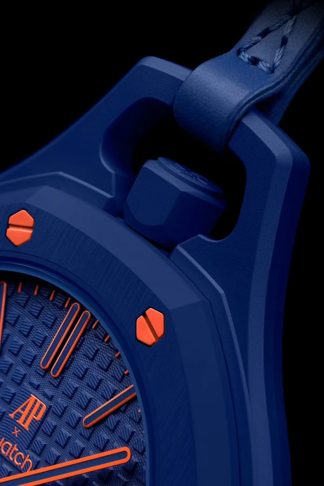 Audemars Piguet ve Swatch iş birliğinden doğan Royal Pop cep saatinin yakın çekiminde, dokulu mavi kadranı üzerinde parlak turuncu indeksler ve akrep yelkovanlar ile mavi kurma kolu ve kasanın köşeli hatları belirginleşiyor.