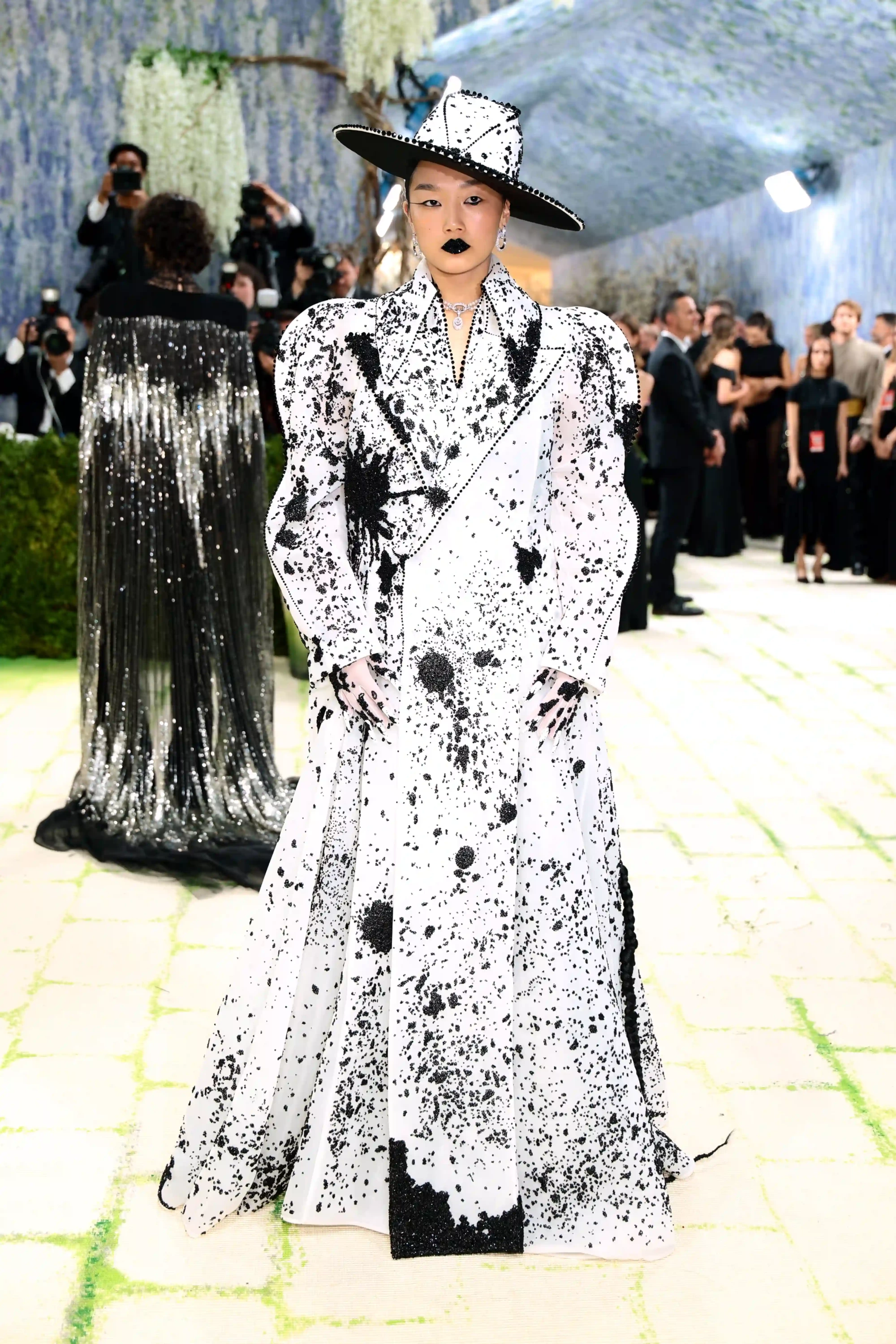 Met Gala 2026 kırmızı halısında, beyaz zemin üzerine dağınık siyah boya sıçramaları ve payet detaylarıyla bezeli, kabarık omuzlu, uzun bir ceket ve uyumlu geniş kenarlı şapkasıyla, koyu renk ruju ve keskin göz makyajıyla iddialı bir kostüm sanatı yorumu sergileyen genç bir kadın tam boy poz veriyor.
