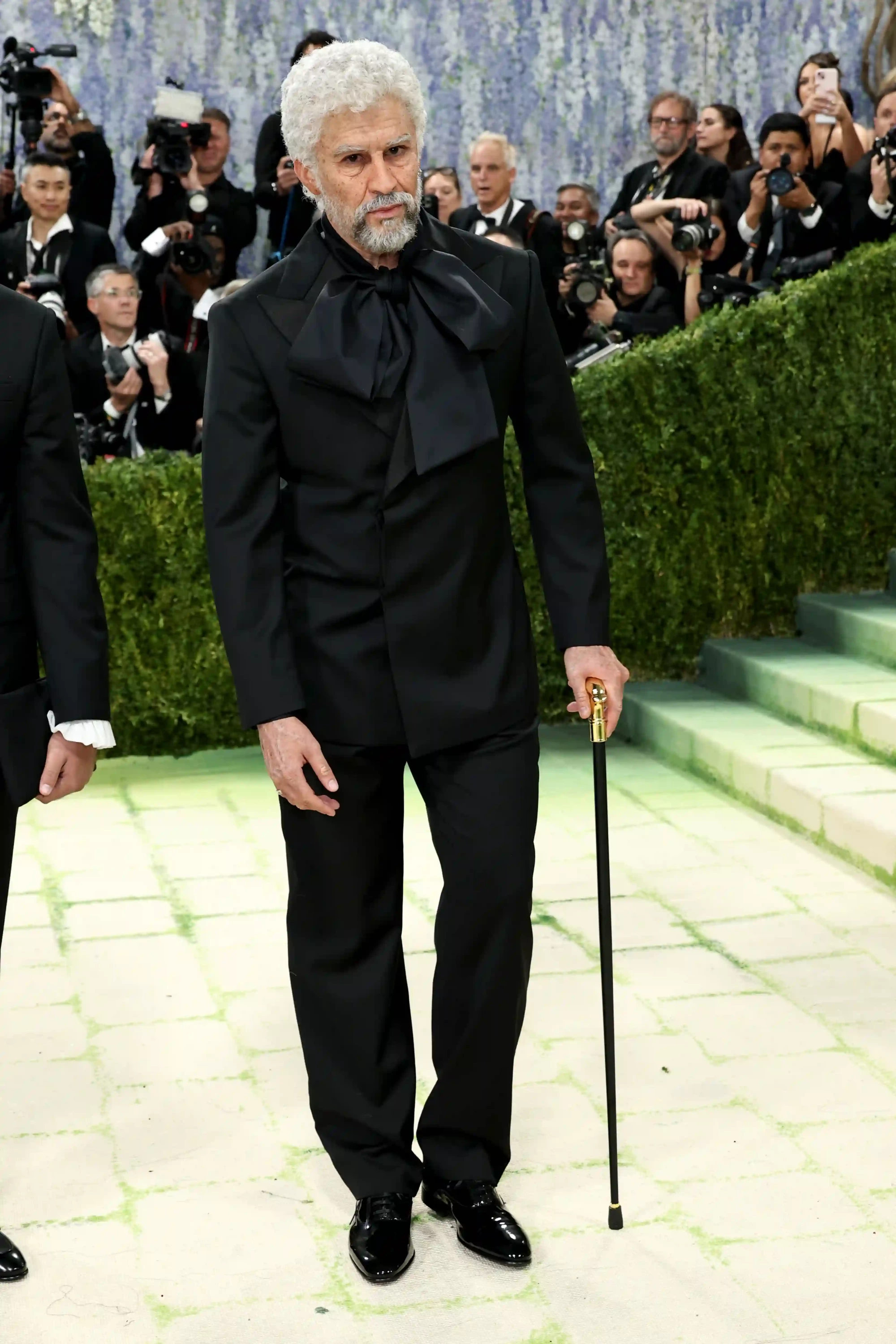 Met Gala 2026'da beyaz kıvırcık saçları ve beyaz sakallarıyla dikkat çeken asil görünümlü bir beyefendi, boynundaki büyük ve kabarık siyah fiyonklu takım elbisesi ve altın başlı bastonuyla yeşil detaylarla bezeli yolda zarifçe duruyor. Arka planda etkinliğin kalabalığı ve fotoğrafçılar flu bir şekilde seçiliyor, bu görünüm moda ve sanat temasına uygun özgün bir tarz sergiliyor.