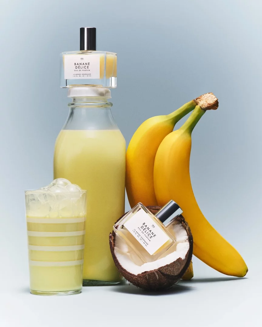 Açık mavi bir arka plan üzerinde, Banane Délice Eau de Parfum şişeleri, taze sarı muzlar, yarım bir hindistan cevizi ve buzlu muzlu içecek dolu çizgili bir bardakla birlikte göz alıcı bir şekilde düzenlenmiş; bir parfüm şişesi uzun cam muzlu içecek şişesinin kapağında dururken, diğeri kırık hindistan cevizinin içinden dışarıya doğru eğik duruyor.