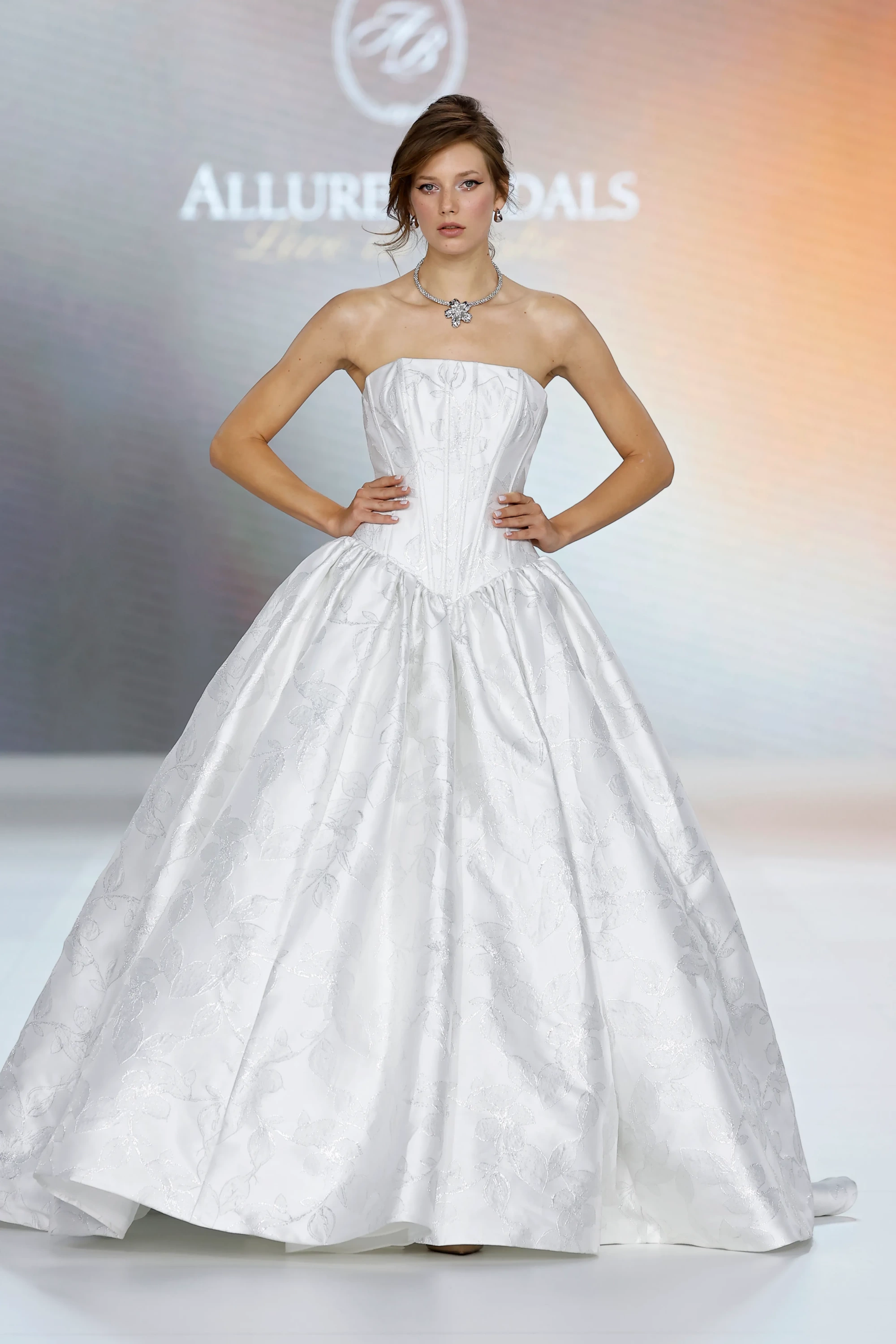 Allure Bridals İlkbahar 2027 Gelinlik Koleksiyonu'ndan, beyaz, omuzları açık straplez bir gelinlik giyen model, yapılandırılmış korsesi ve tüm etek boyunca uzanan zarif yaprak desenleriyle klasik prenses siluetini modern bir dokunuşla yorumluyor; elleri belinde poz veren modelin şıklığını, boynundaki büyük taşlı kolye ve küpeleri tamamlıyor.