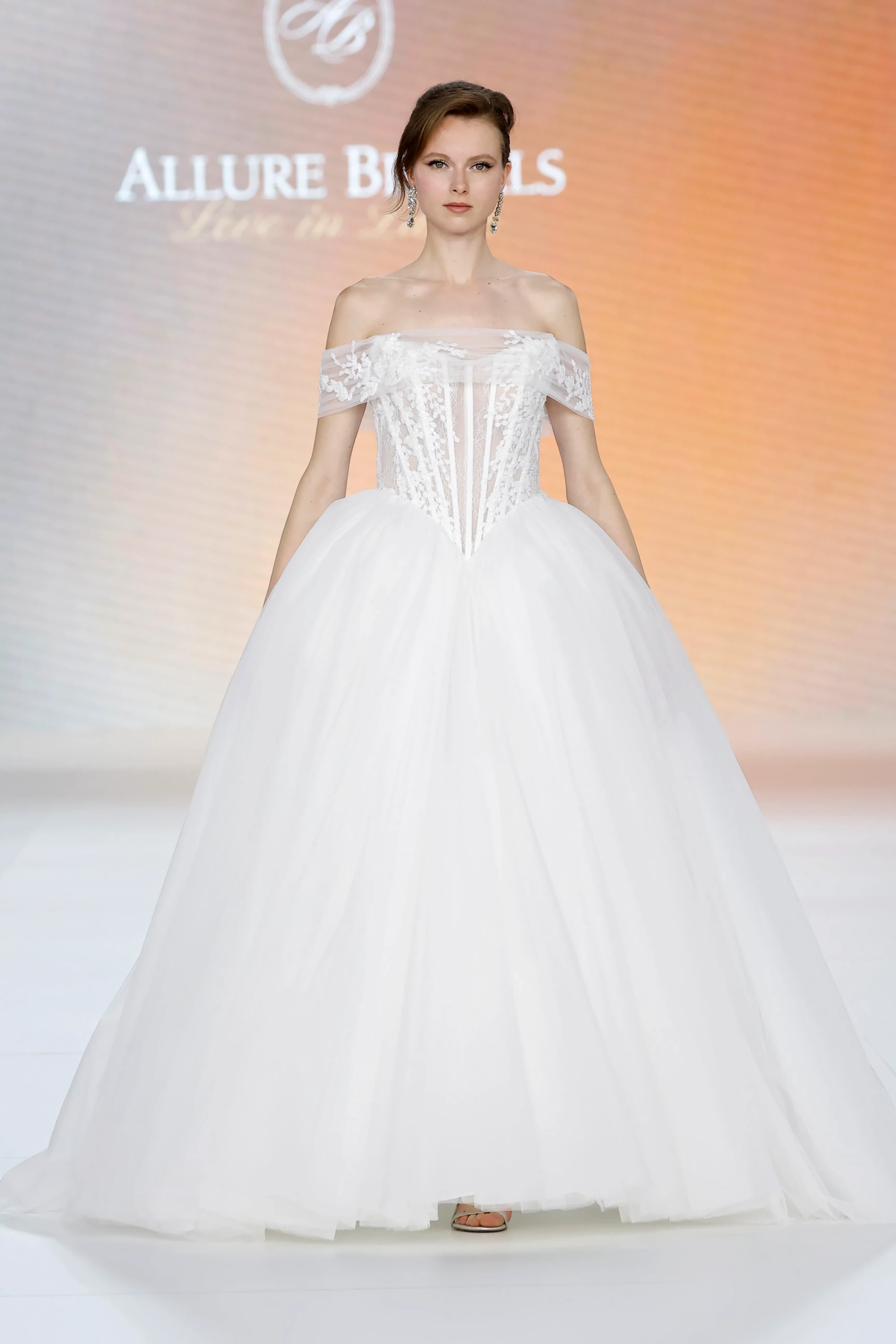 Allure Bridals İlkbahar 2027 Gelinlik Koleksiyonu'ndan, omuzları açıkta bırakan dantel işlemeli korsajı ve katmanlı tül ile oluşturulmuş, zarif ve hacimli prenses eteğiyle klasik çizgiyi modern bir şıklıkla buluşturan beyaz bir gelinlik giyen model podyumda duruyor.