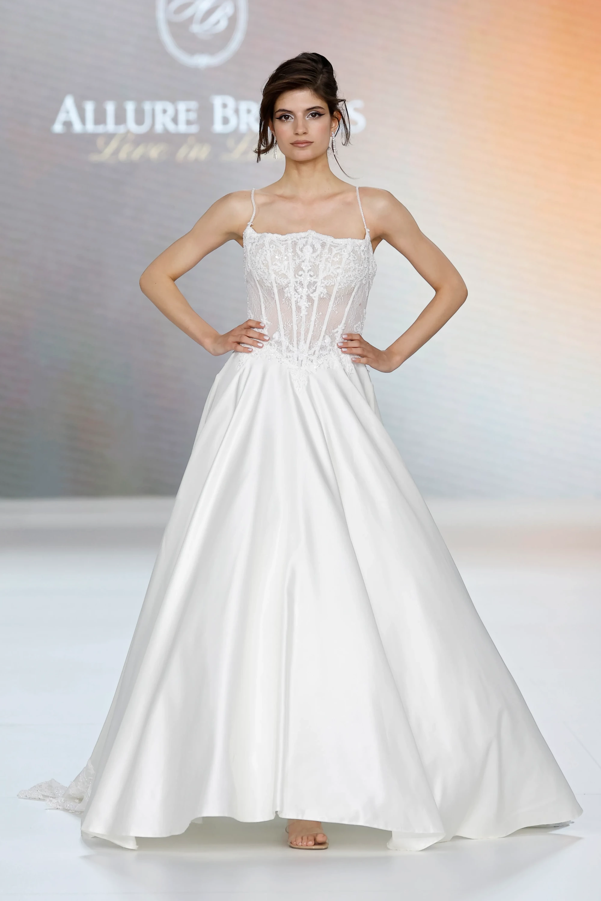 Bir podyumda elleri belinde poz veren model, transparan korseli ve romantik dantel işlemeli üst kısmı ile parlak, hacimli, yere kadar uzanan A kesim eteği olan zarif bir Allure Bridals İlkbahar 2027 gelinlik giyiyor.