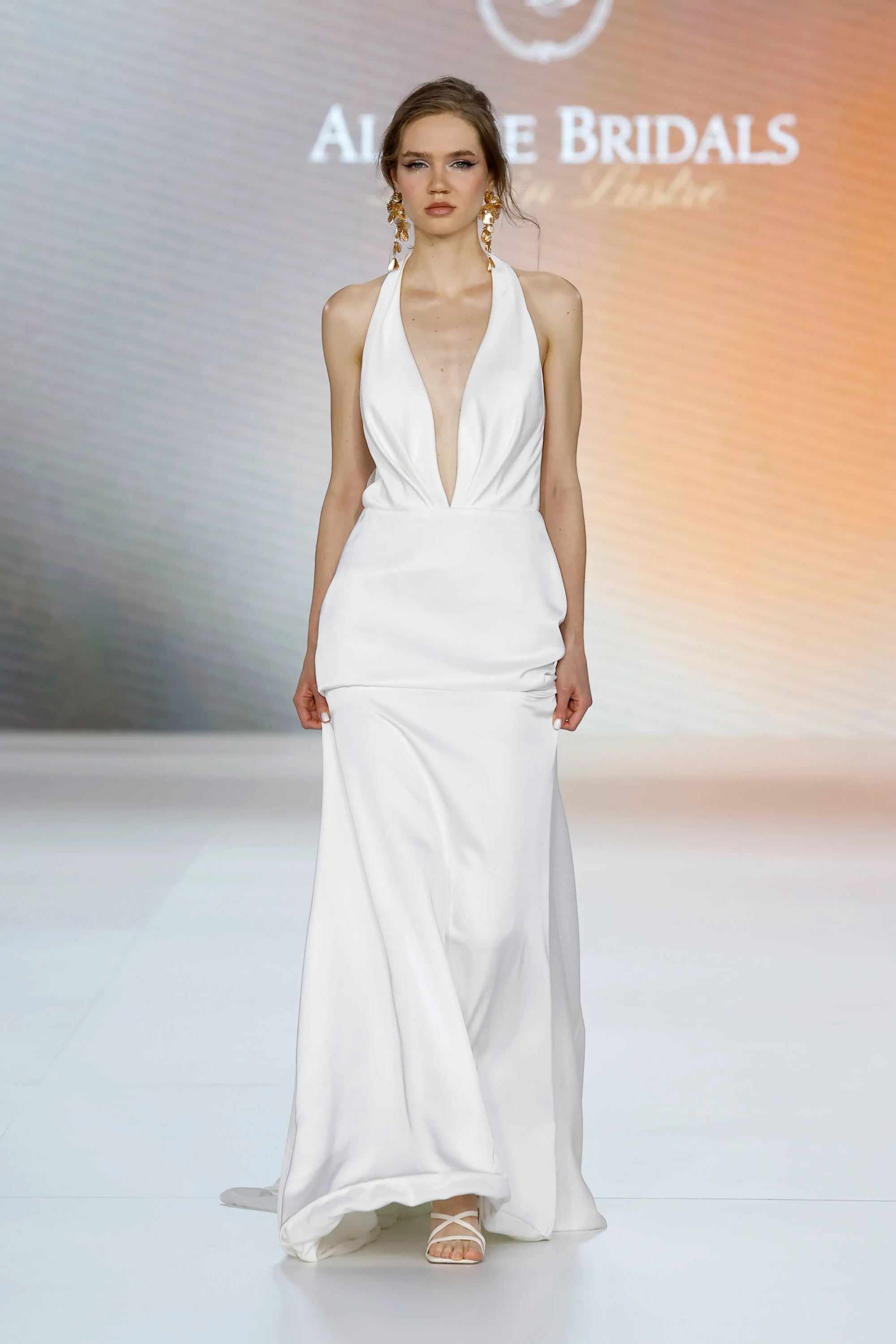 Allure Bridals İlkbahar 2027 Gelinlik Koleksiyonu defilesinde podyumda yürüyen bir model, derin V yakalı, vücuda oturan üst kısmı ve akışkan, sade etekli beyaz bir gelinlikle modern ve zamansız bir zarafet sunuyor, görünümünü dikkat çekici büyük altın küpelerle tamamlıyor.