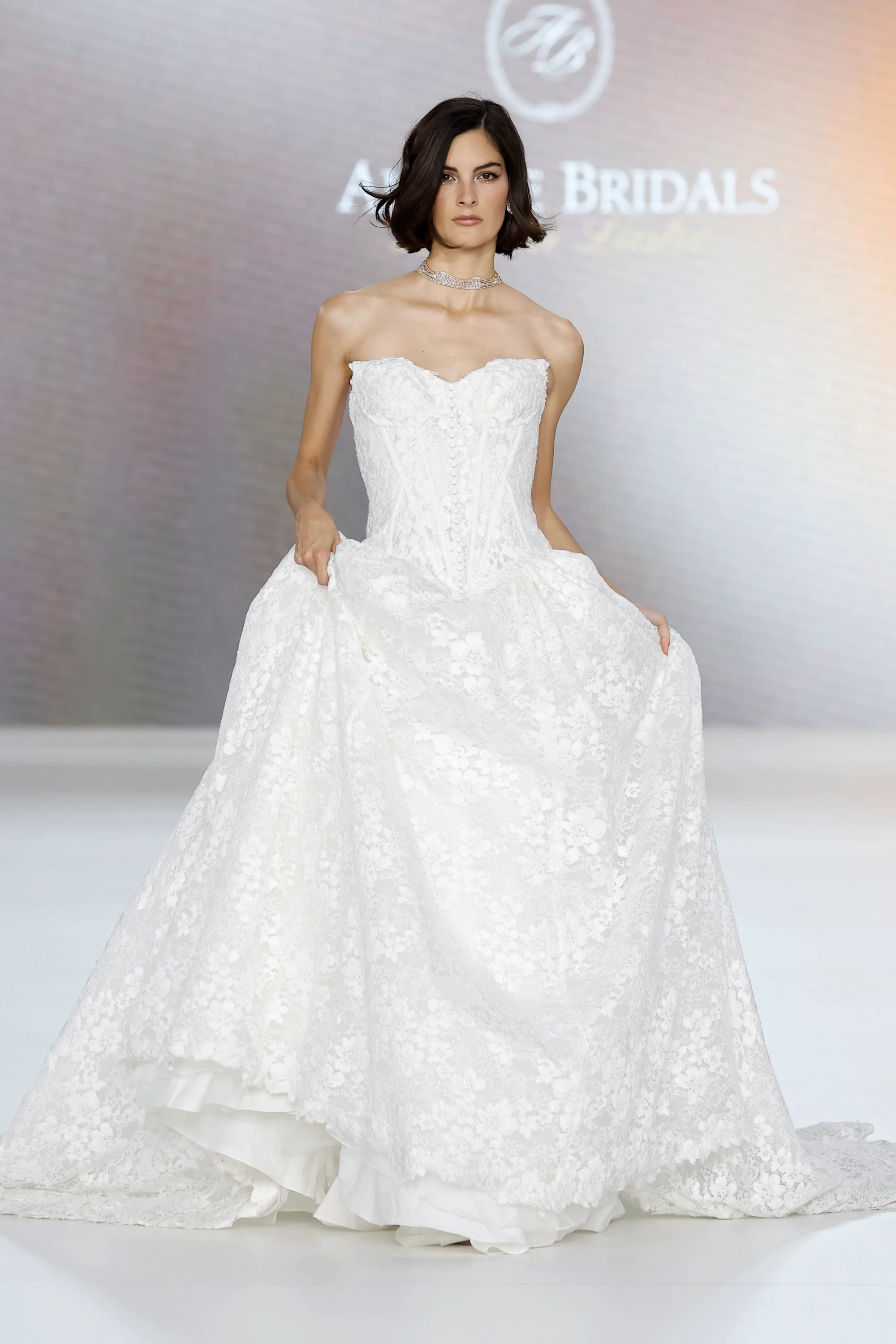 Allure Bridals İlkbahar 2027 Gelinlik Koleksiyonu'ndan, pistte kısa bob saçlı, taşlı choker kolyesiyle zarif bir model, narin işlemeli yapılandırılmış dantel korsajlı ve hacimli, katmanlı etekli göz alıcı straplez beyaz prenses gelinliğini sergiliyor.