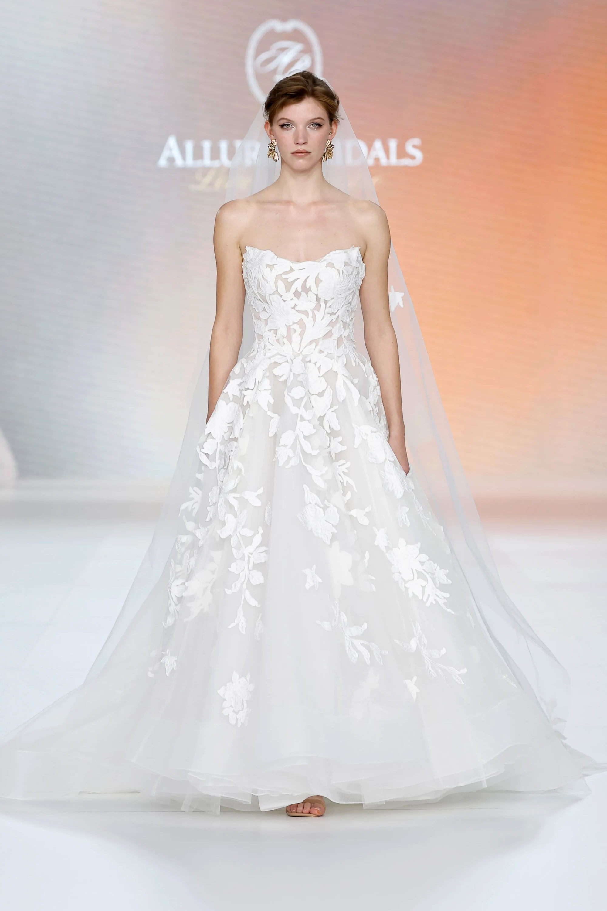 Allure Bridals İlkbahar 2027 Gelinlik Koleksiyonu'nun modern zarafetini yansıtan bu fotoğrafta, straplez kesime sahip, tül üzerine işlenmiş beyaz çiçek aplikeleriyle romantik bir hava kazanan geniş etekli gelinlik ve uzun tül duvağıyla podyumda duran zarif bir model görülüyor.