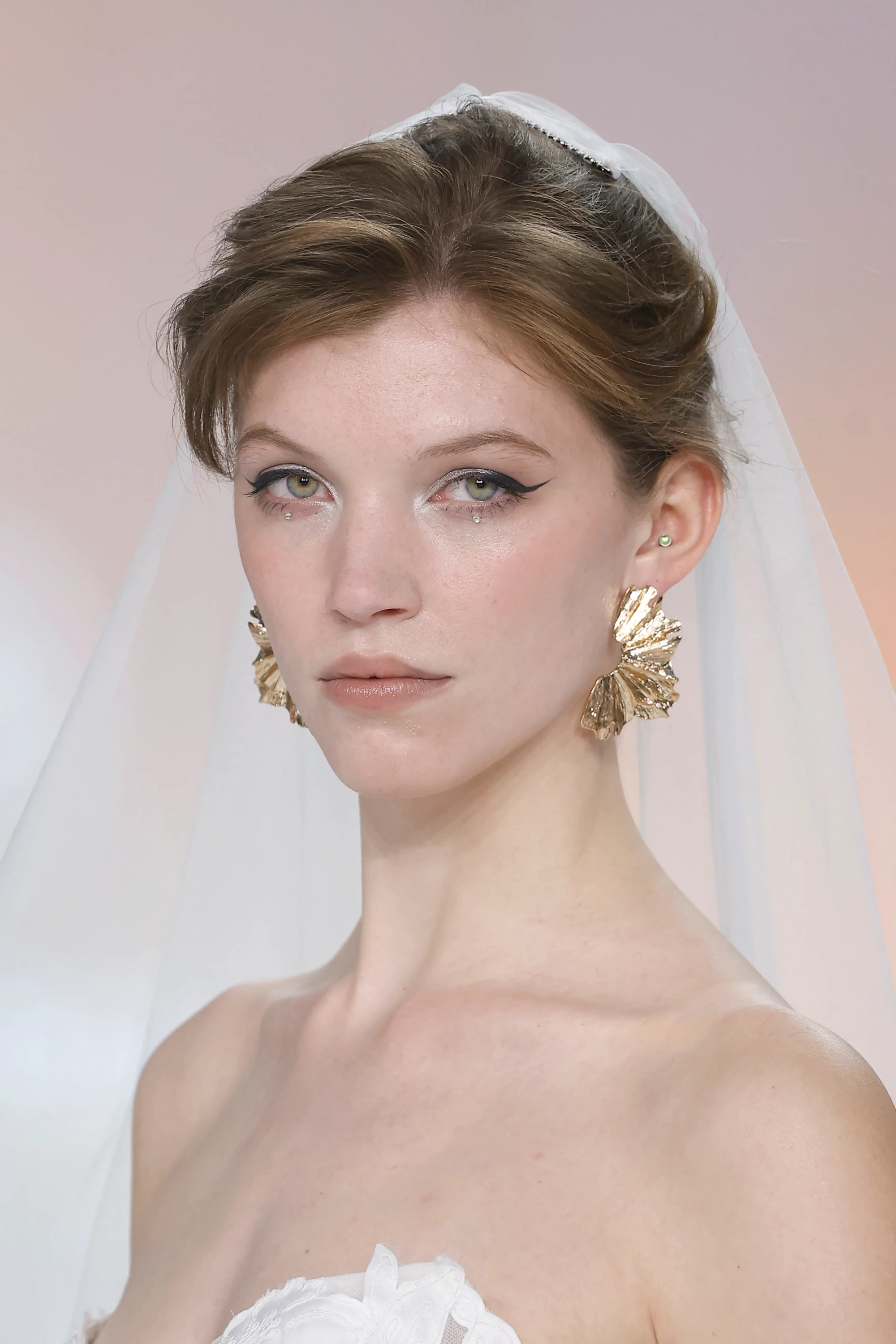 Straplez gelinlik giyen bir modelin yakından çekiminde, belirgin eyeliner ve göz altı taşlarıyla vurgulanan gözleri, şık bir topuz ve tül duvak, Allure Bridals İlkbahar 2027 Gelinlik Koleksiyonu'nun modern zarafetini yansıtan dalgalı altın küpelerle tamamlanıyor.
