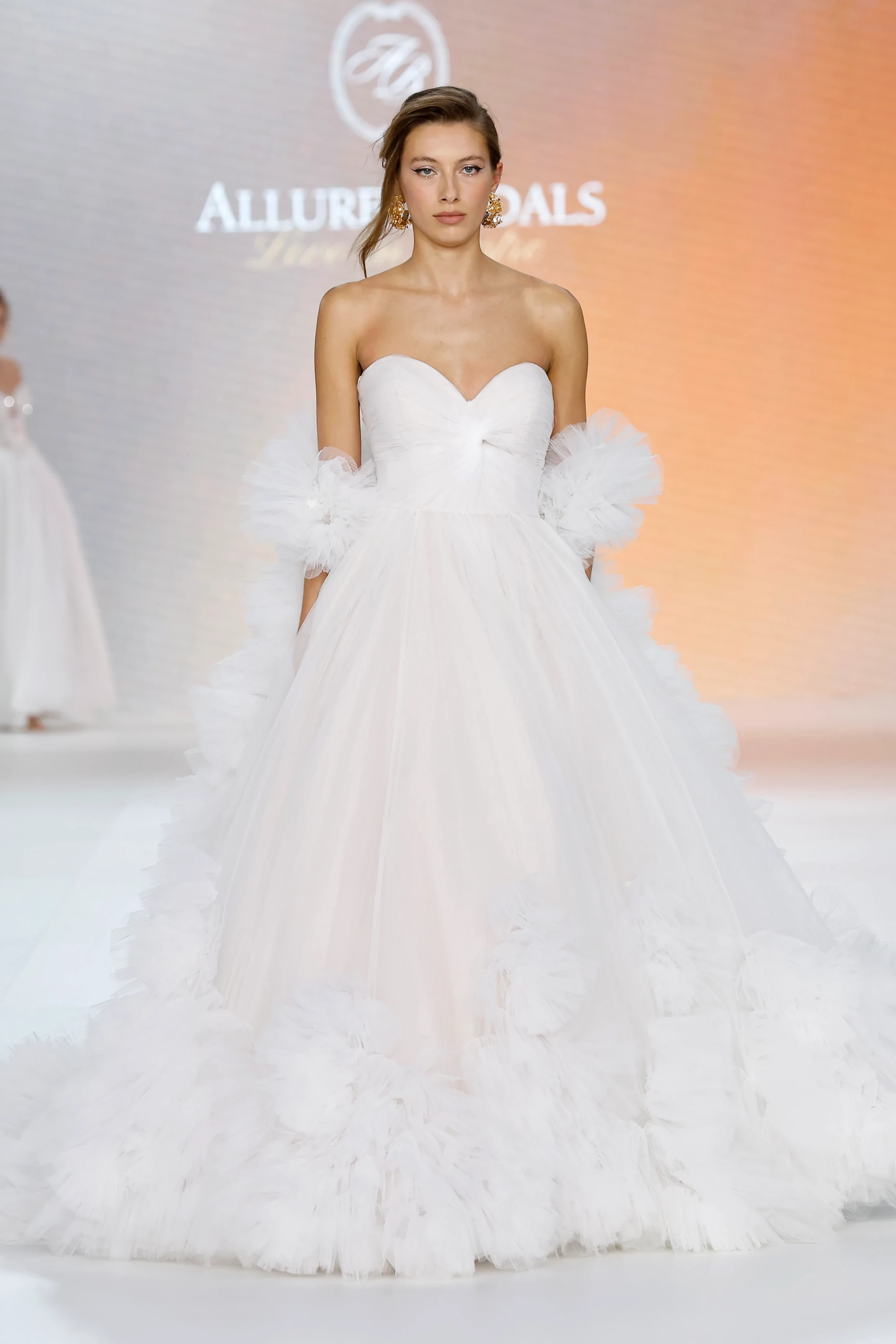 Runway üzerinde, straplez kalp yaka beyaz gelinlik giyen bir model, kabarık tül katmanlarından oluşan hacimli eteği ve omuzlarındaki fırfırlı tül detayları ile Allure Bridals İlkbahar 2027 Gelinlik Koleksiyonu'ndan modern ve zarif bir görünüm sergiliyor.