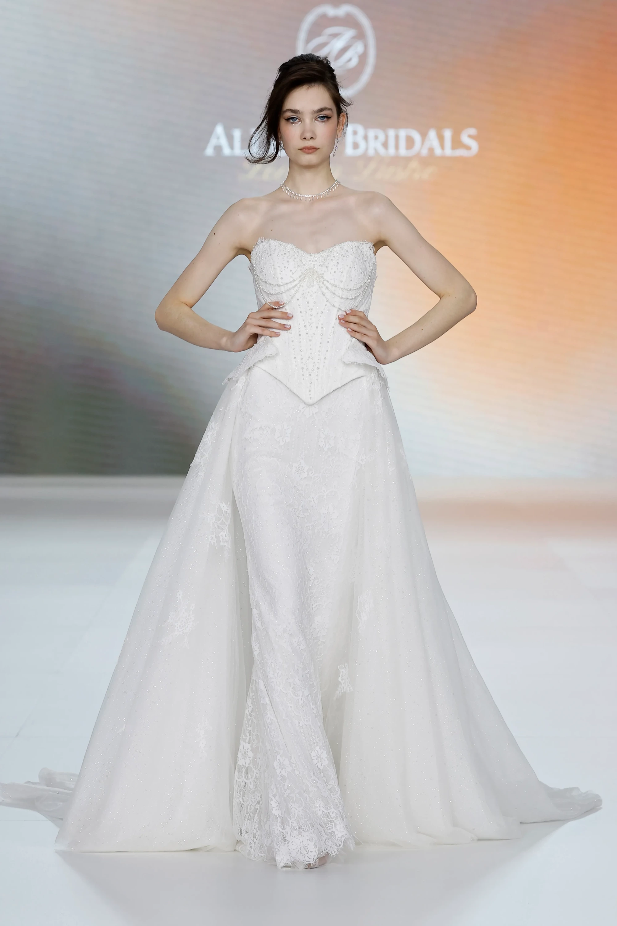 Bir model, Allure Bridals yazılı podyumda, incilerle bezeli, yapılandırılmış straplez korsajı, belden açılan peplum detayı, dantelli dar iç elbisesi ve ışıltılı, katmanlı, hacimli tül dış eteğiyle Allure Bridals İlkbahar 2027 Gelinlik Koleksiyonu'ndan modern bir gelinlik sergiliyor.