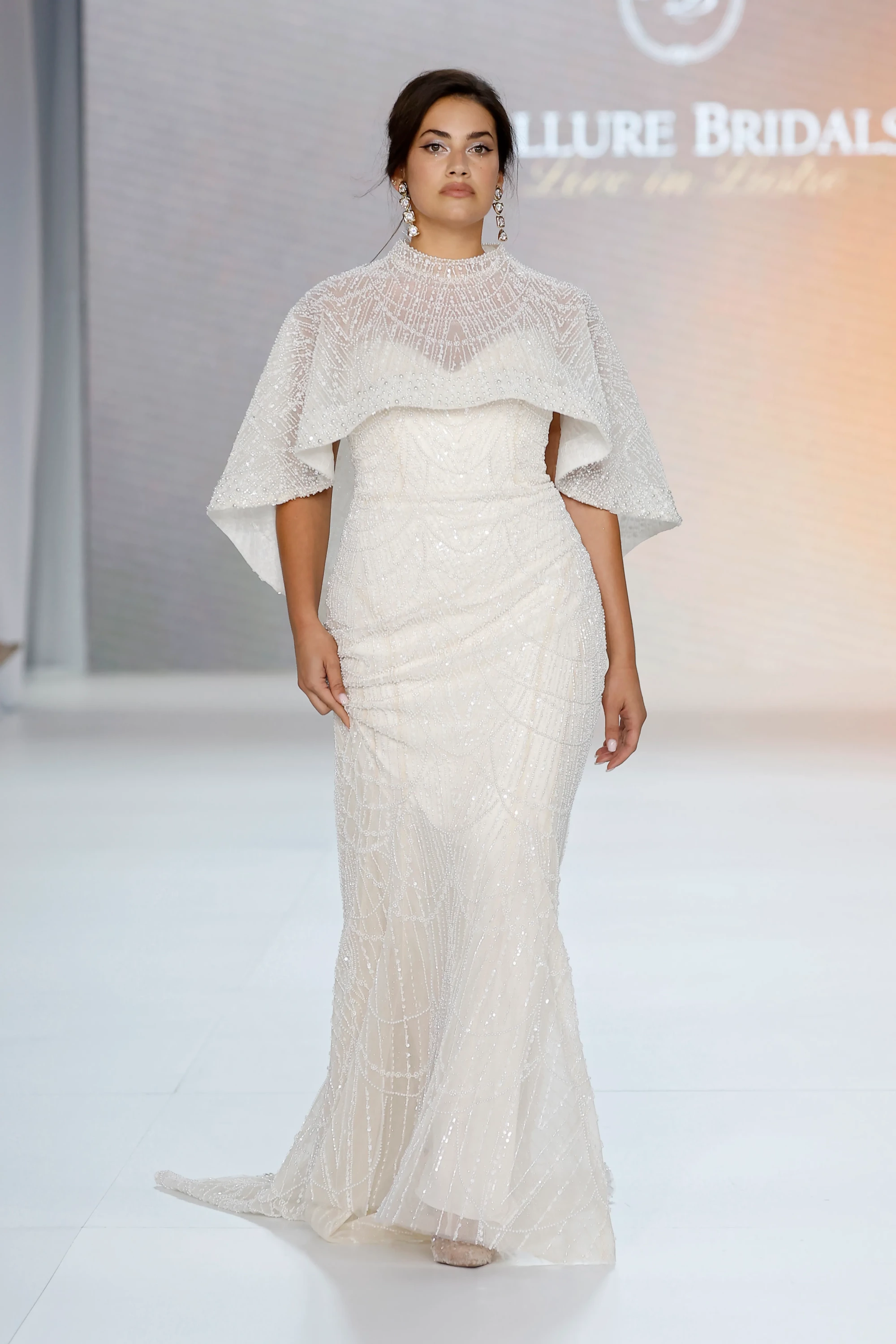 Allure Bridals İlkbahar 2027 Gelinlik Koleksiyonu'ndan, vücuda oturan, boyundan yüksek şeffaf pelerinli ve tamamı işlemeli straplez gelinliğiyle podyumda yürüyen bir model, modern zarafeti ve katmanlı şıklığı vurguluyor.