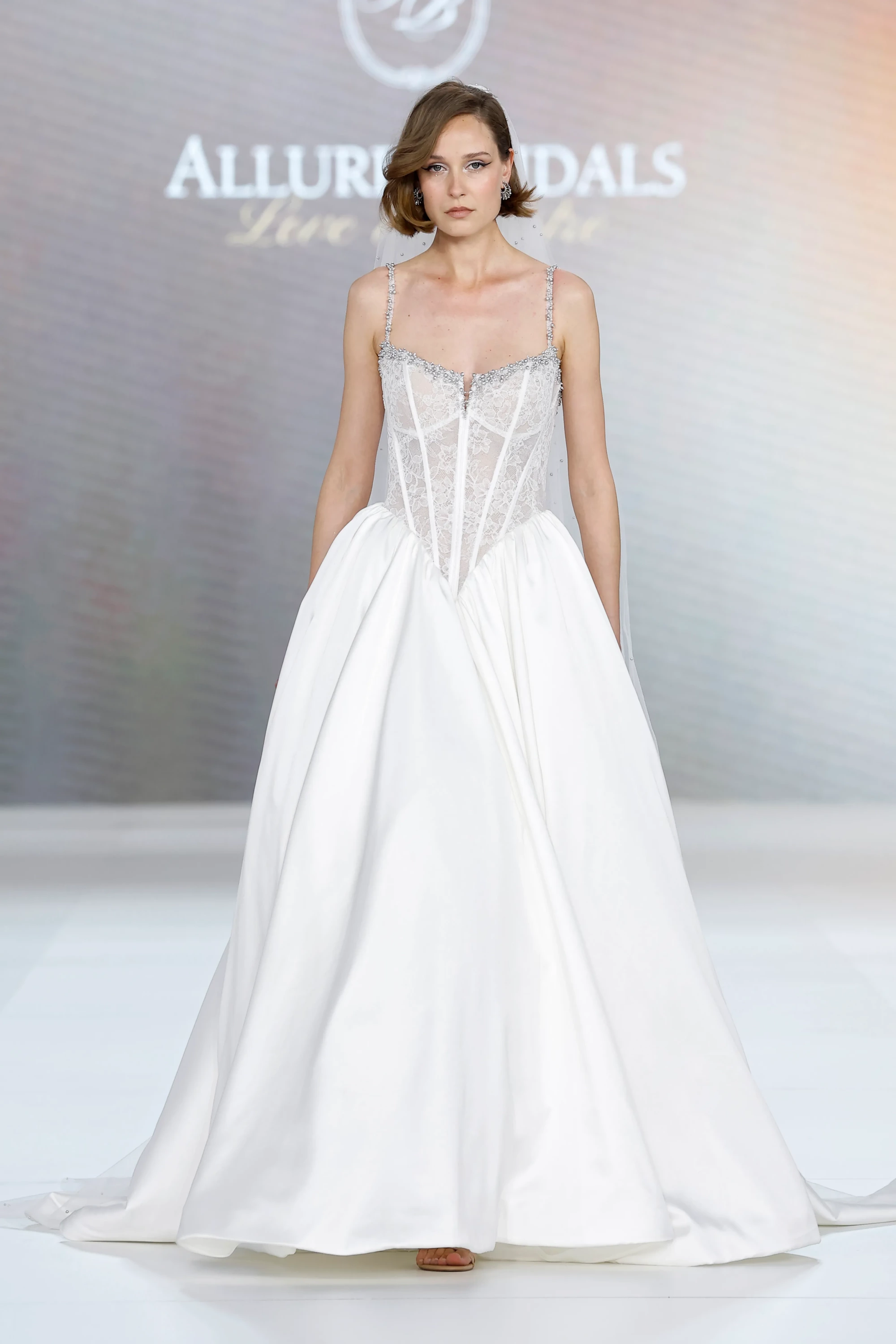 Model, podyumda Allure Bridals İlkbahar 2027 Gelinlik Koleksiyonu'ndan, şeffaf dantel korse ve boncuk işlemeli zarif askılarla tamamlanan, hacimli saten eteği ve gizli cepleriyle modern ve sofistike bir gelinlik sunuyor.