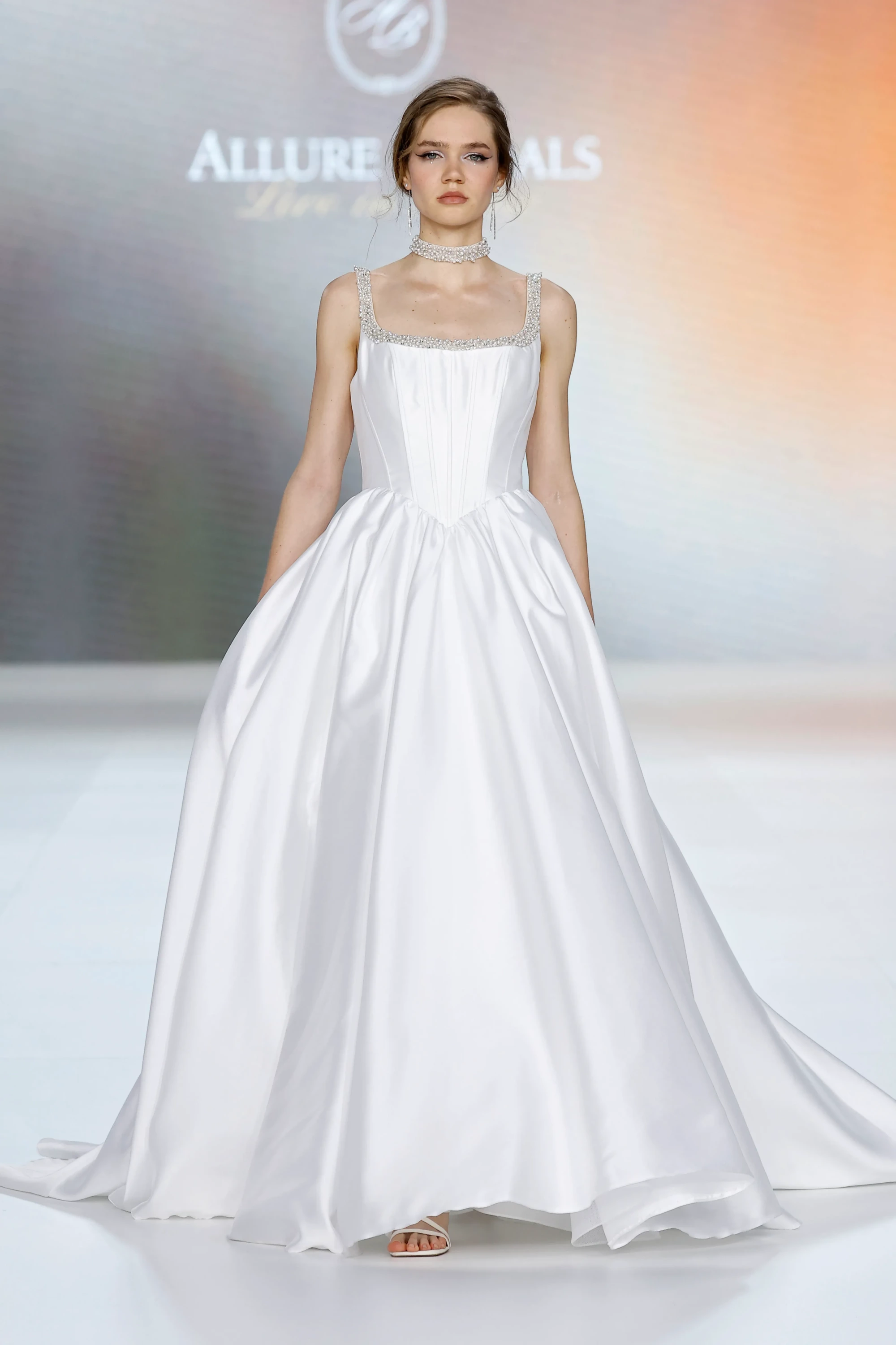 Allure Bridals İlkbahar 2027 Gelinlik Koleksiyonu'ndan bir model, boncuk işlemeli kare yakalı ve kalın askılı, yapılandırılmış korsajlı, belden aşağıya doğru hacimli ve yere kadar uzanan saten beyaz bir prenses silüet gelinlik giyerek, taşlı choker kolyesi, sallantılı küpeleri ve göz alıcı makyajıyla podyumda modern bir zarafet sunuyor.