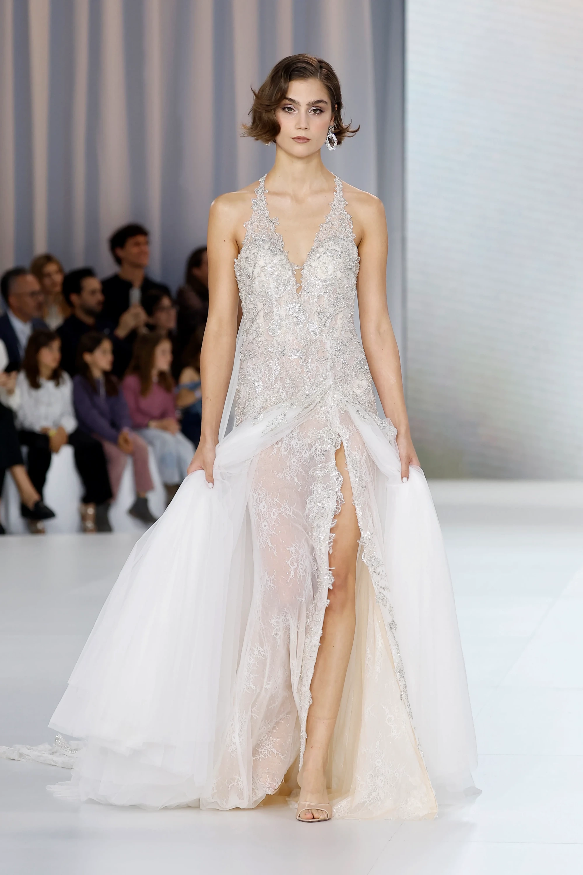 Allure Bridals İlkbahar 2027 Gelinlik Koleksiyonu'ndan modern bir tasarımı sergileyen bir model, podyumda derin V yakalı, ışıltılı dantel işlemeli korseli ve yüksek yırtmaçlı transparan iç eteği, iki yandan tuttuğu akışkan hacimli tül dış eteğiyle sofistike ve katmanlı bir görünüm sunuyor.