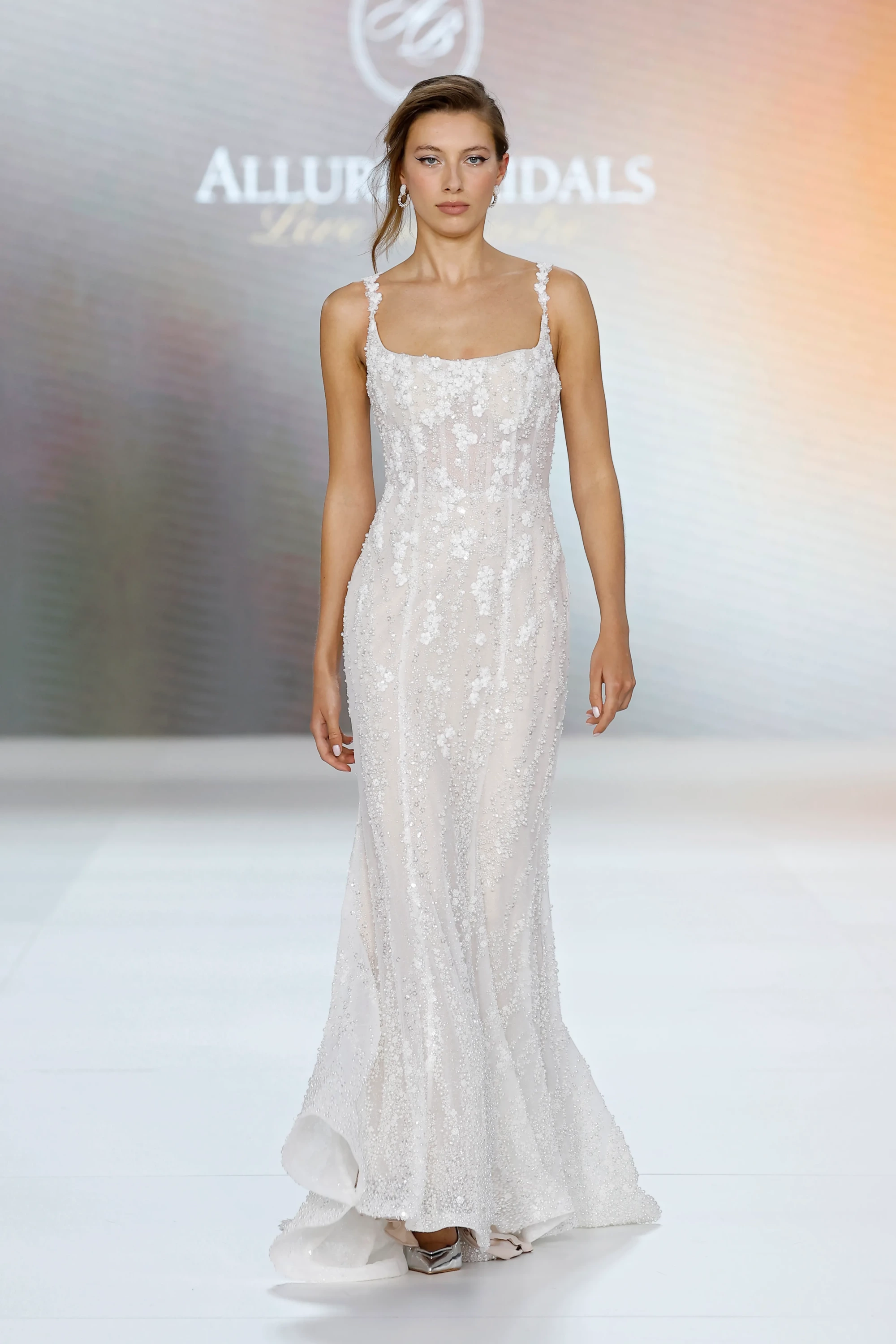 Allure Bridals İlkbahar 2027 Gelinlik Koleksiyonu'ndan, podyumda duran bir model, ince askılı, bedeni saran ve etek kısmı hafifçe açılan, tamamı ışıltılı boncuklar ve zarif çiçek işlemeleriyle süslenmiş modern ve akışkan formda bir gelinlik sergiliyor.
