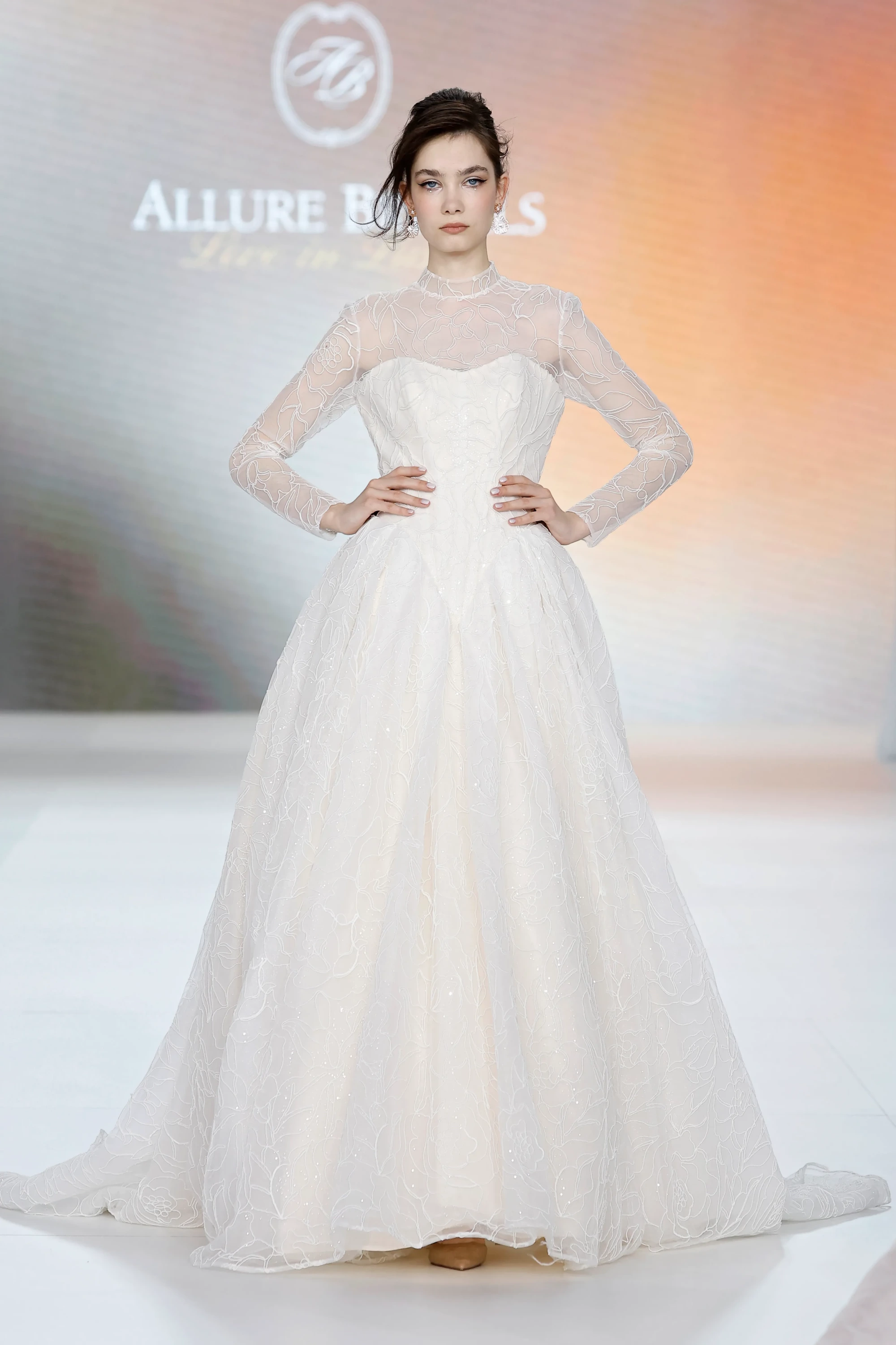 Allure Bridals İlkbahar 2027 Gelinlik Koleksiyonu'ndan, zarif çiçek işlemeli transparan uzun kollu tül yaka detayıyla modern bir dokunuş kazanan, hacimli eteği ve straplez korsesiyle zamansız bir şıklık sunan gelinlik giymiş bir manken podyumda duruyor.