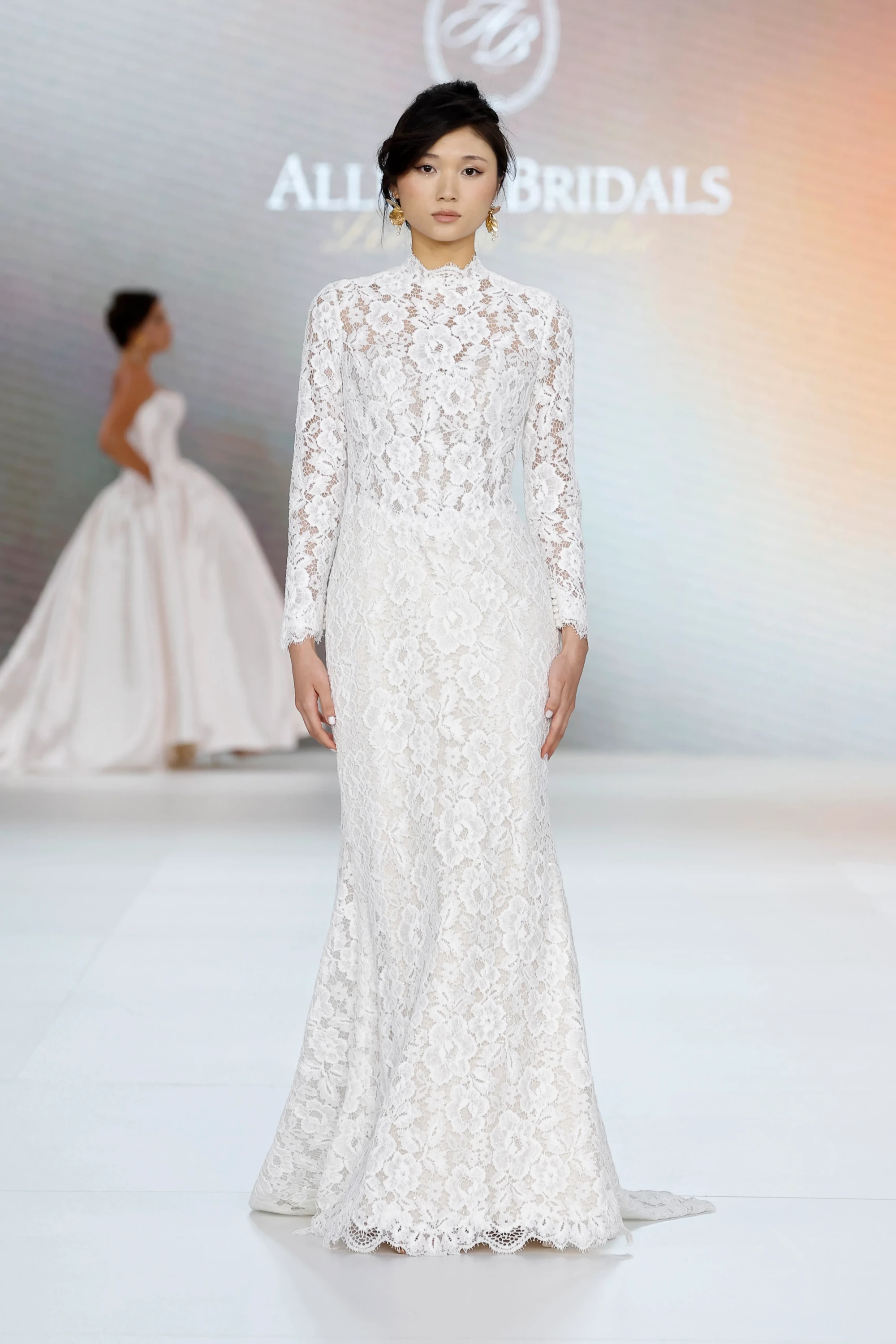 Allure Bridals İlkbahar 2027 Gelinlik Koleksiyonu'ndan, podyumda duran bir model, modern bir yorumla tasarlanmış, vücuda oturan, uzun kollu, dik yakalı ve baştan aşağı zarif çiçek desenli beyaz dantel gelinlik giyiyor.