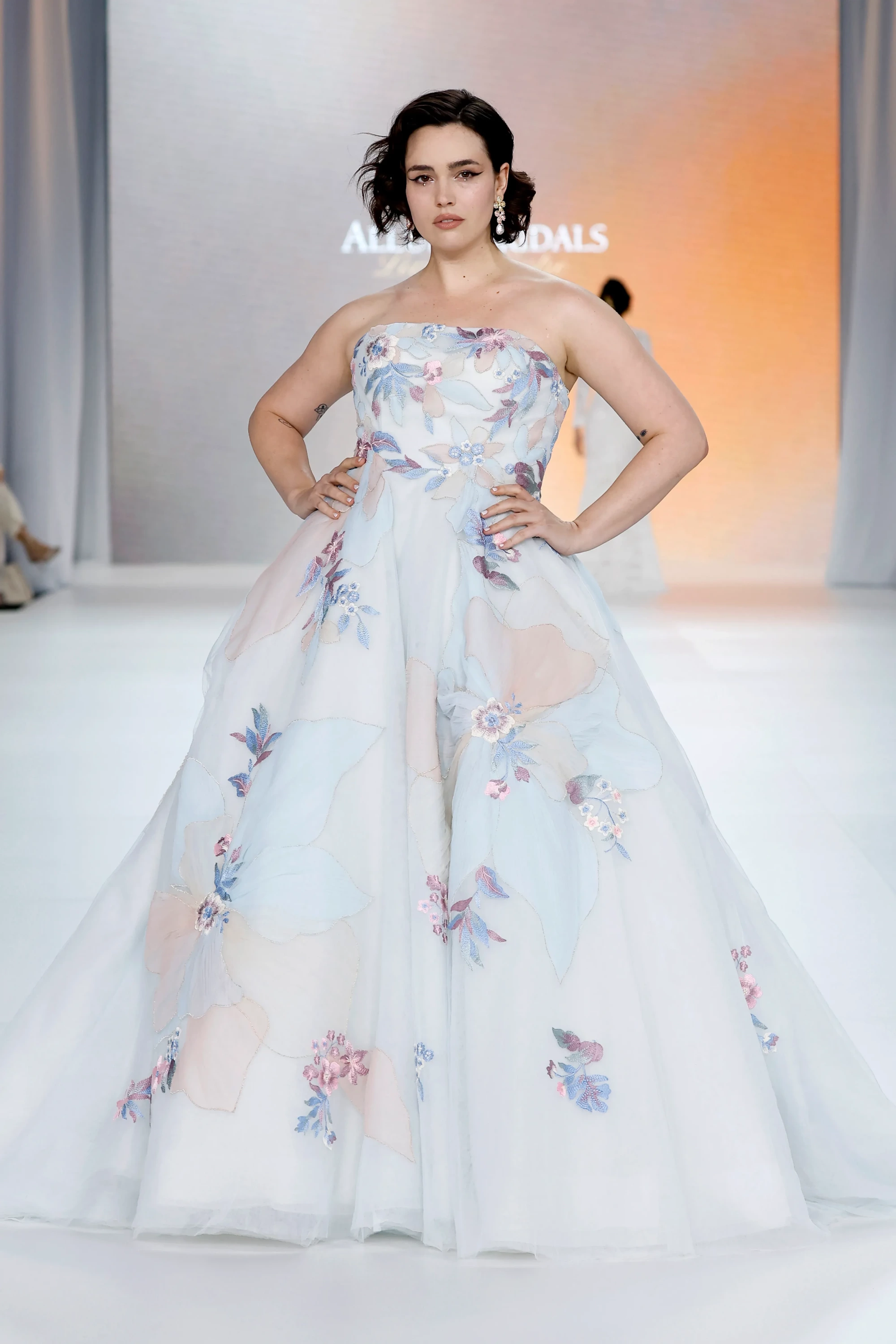 Allure Bridals İlkbahar 2027 Gelinlik Koleksiyonu'ndan, straplez kesimli, hacimli tül eteği pastel tonlarda rengarenk çiçek işlemeleriyle bezeli gelinliği podyumda sergileyen, kısa saçlı, belirgin göz makyajlı bir model modern ve rafine bir duruş sergiliyor.