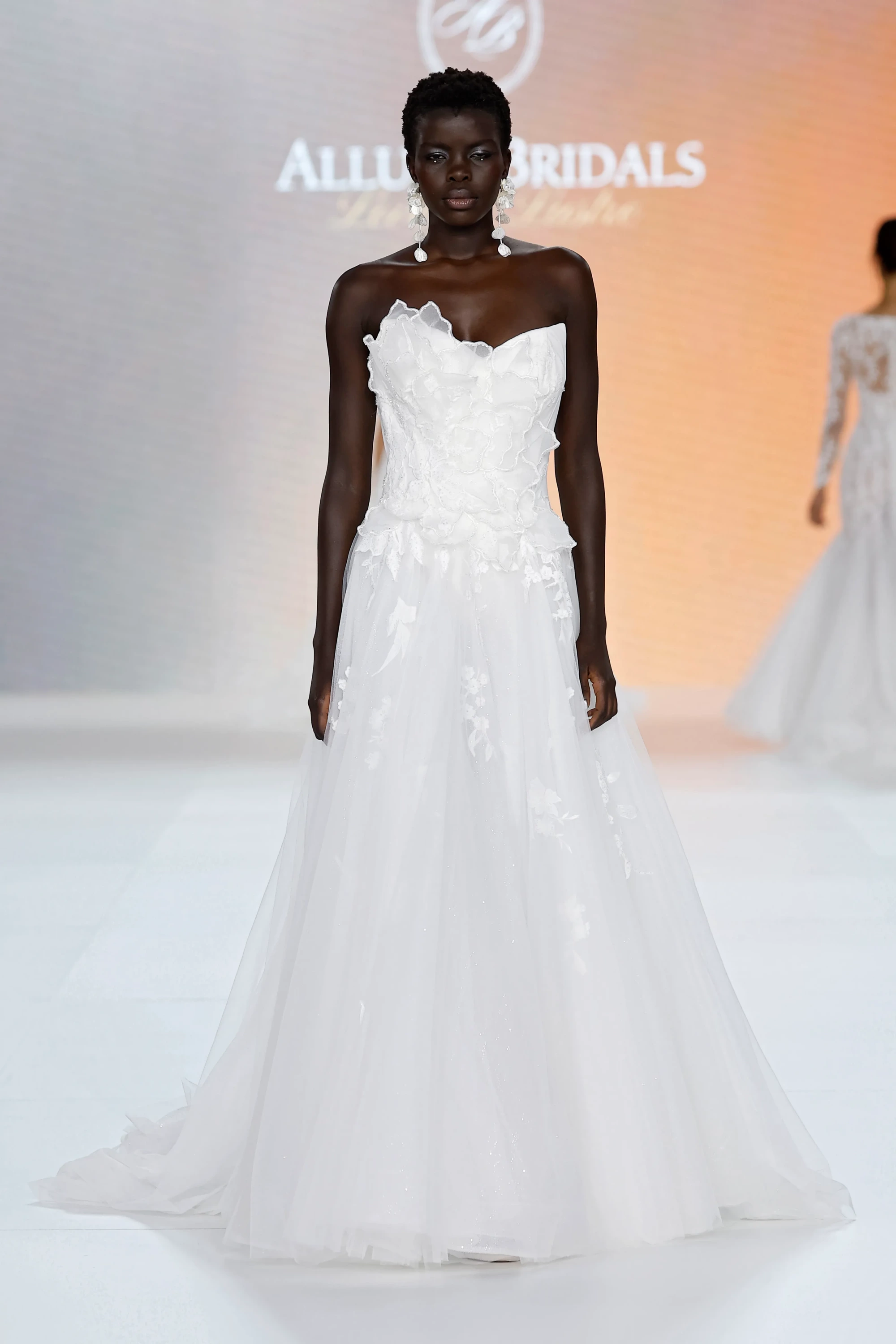 Allure Bridals İlkbahar 2027 Gelinlik Koleksiyonu'ndan, straplez kesimli, korse kısmında fırfır ve ince işlemelerle zenginleştirilmiş, hacimli katmanlı tül eteğinde hafif parıltılar ve bitkisel dantel detaylar taşıyan beyaz bir gelinlikle podyumda yürüyen model, stilini büyük, şeffaf çiçek formlu küpelerle tamamlayarak klasik zarafete çağdaş bir dokunuş katıyor.