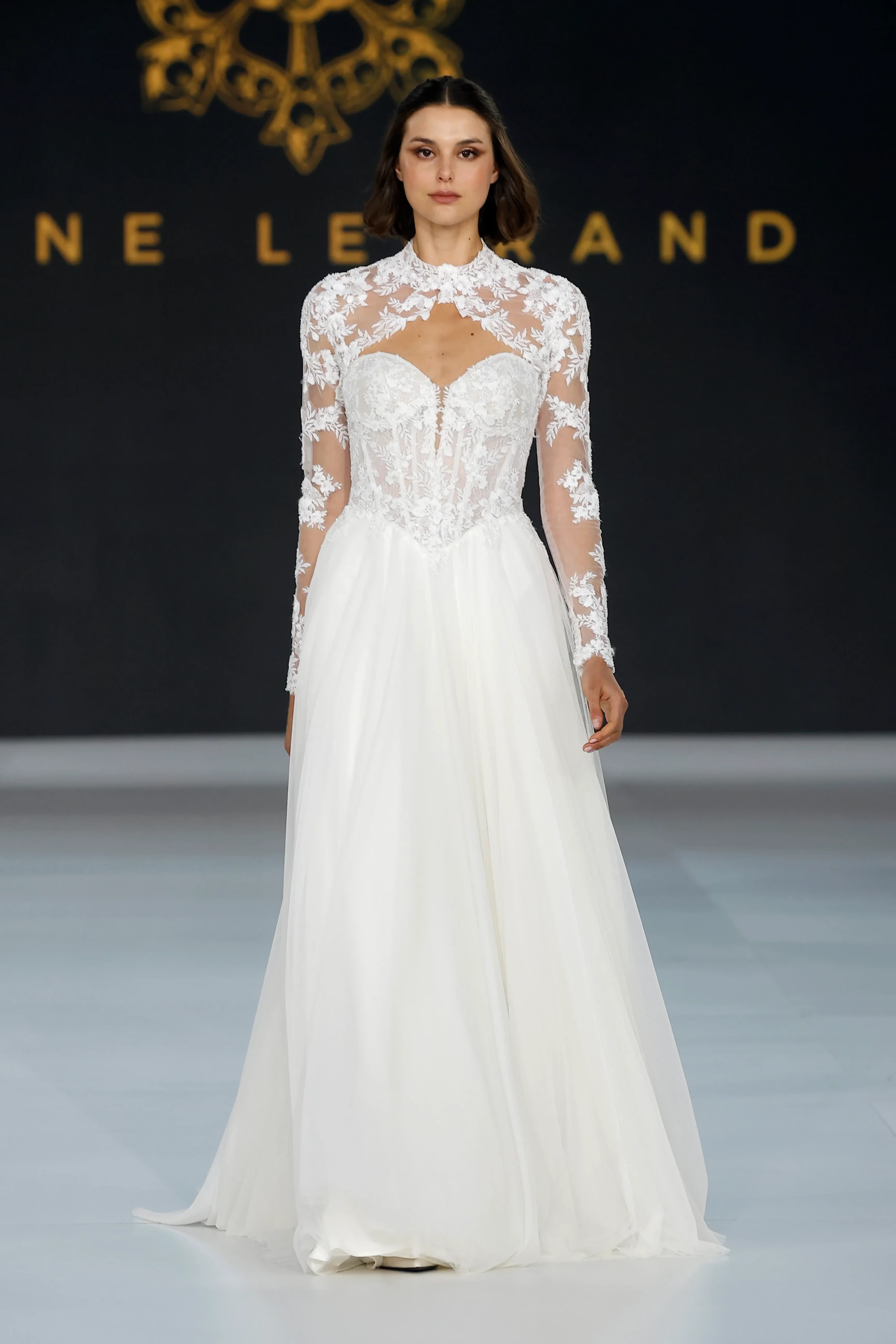 Barcelona Bridal Fashion Week'te podyumda yürüyen bir model, Diane Legrand İlkbahar 2027 Gelinlik Koleksiyonu'ndan yüksek yakalı, uzun kollu, transparan dantel işlemeli korseli üst kısmı ve katmanlı tül eteğiyle zarif bir gelinlik sunuyor.