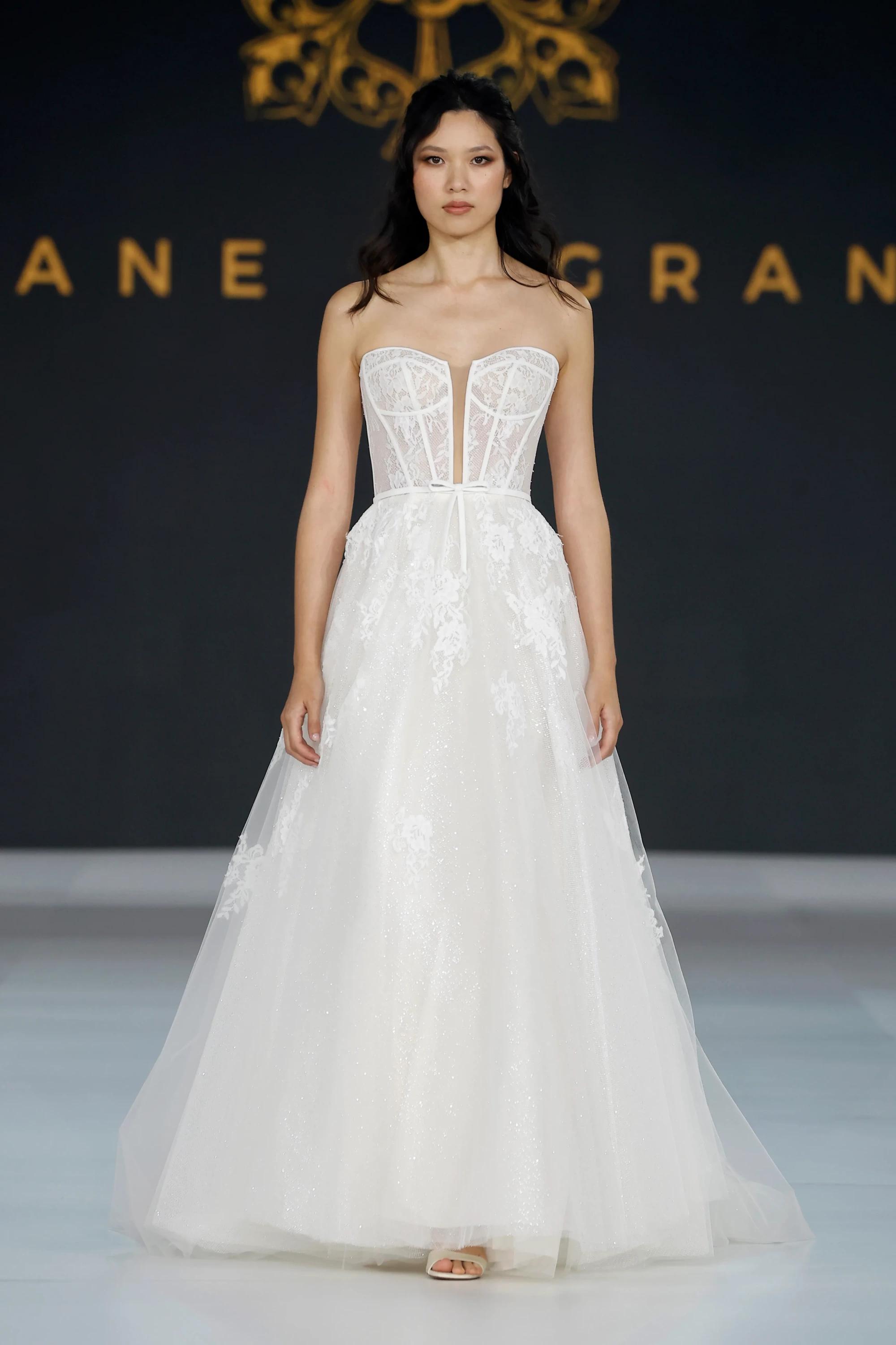 Model, Diane Legrand İlkbahar 2027 Gelinlik Koleksiyonu'ndan straplez, dantel ve şeffaf panellerle detaylandırılmış korseli üst kısmı ve ışıltılı tül katmanlarıyla hacimli etek kısmı olan zarif, beyaz bir gelinlik sergiliyor.