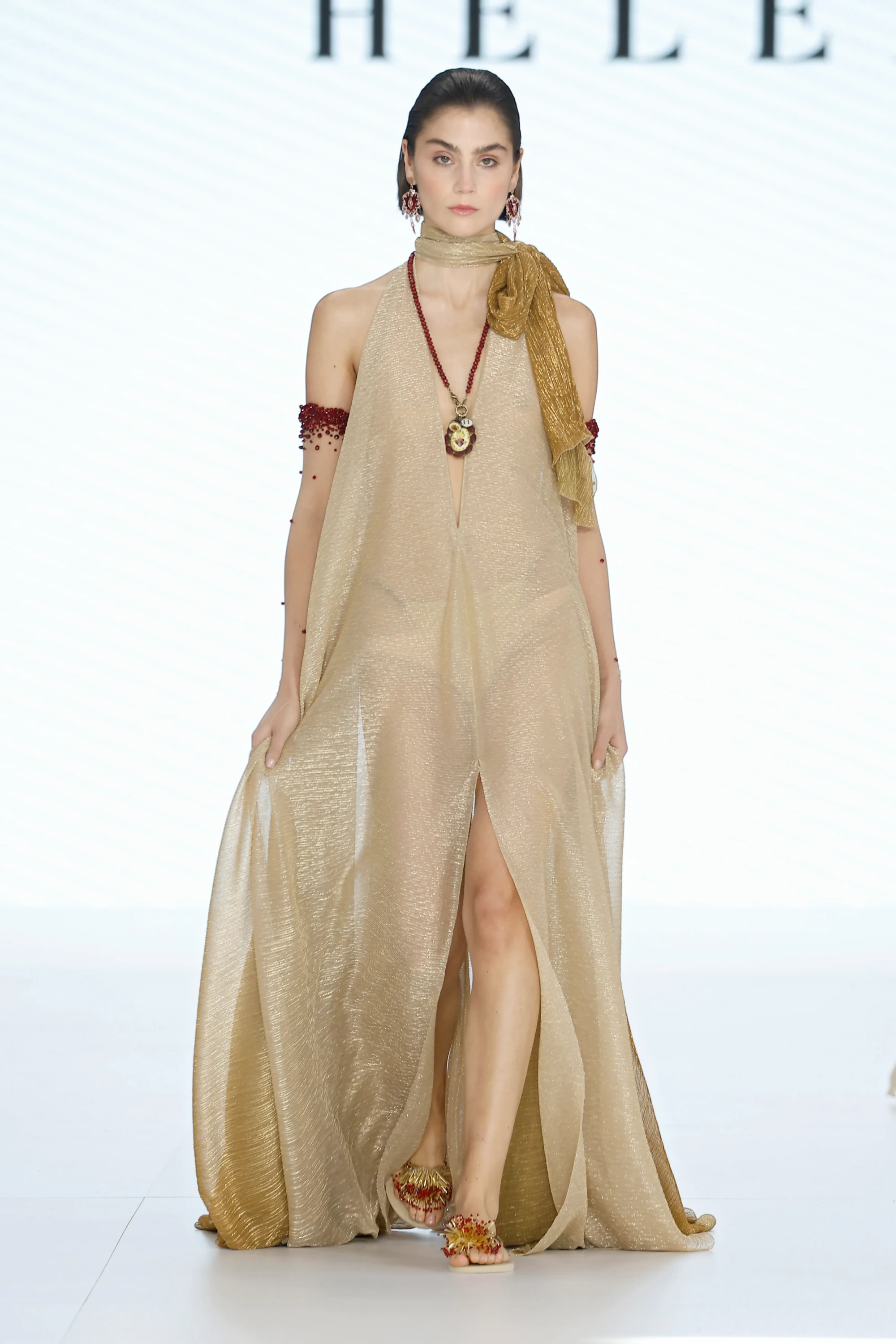 Barcelona Bridal Fashion Week 2026'da podyumda yürüyen Helena İlkbahar 2027 Gelinlik Koleksiyonu'ndan, şeffaf, metalik altın rengi, derin V yakalı ve önü yırtmaçlı elbisesiyle dikkat çeken model, Sacred Heart ikonografisinden ilham alan kırmızı boncuklu aksesuarları ve iddialı sandaletleriyle dramatik bir ifade sergiliyor.