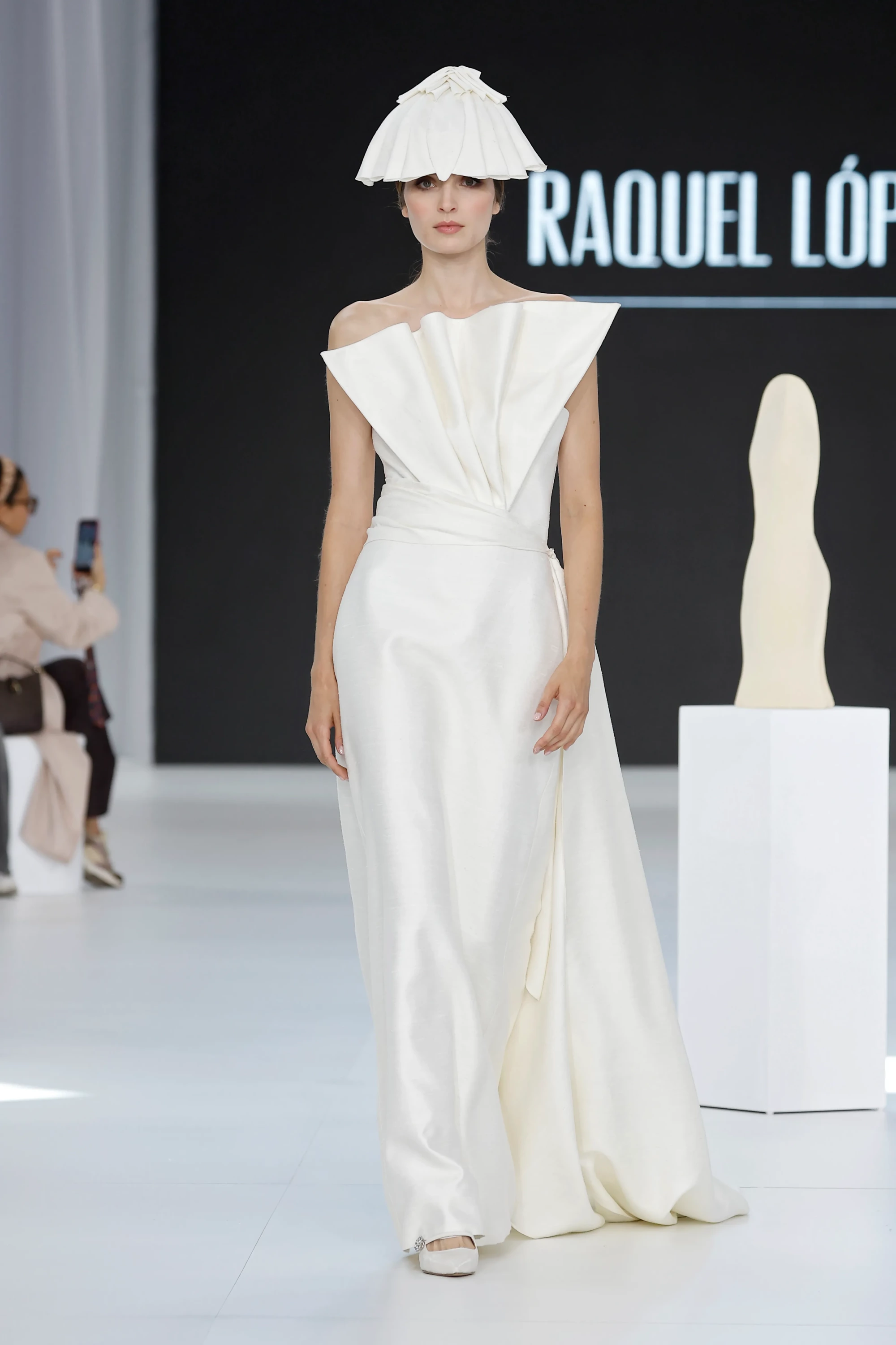 Raquel López İlkbahar 2027 Gelinlik Koleksiyonu'ndan, podyumda duran bir model, heykelsi kıvrımlara sahip straplez üst kısmı ve vücudu zarifçe saran akışkan eteğiyle dikkat çeken fildişi rengi gelinliği ve katmanlı, pilili şapkamsı başlığıyla modern ve yalın bir zarafet sergiliyor.