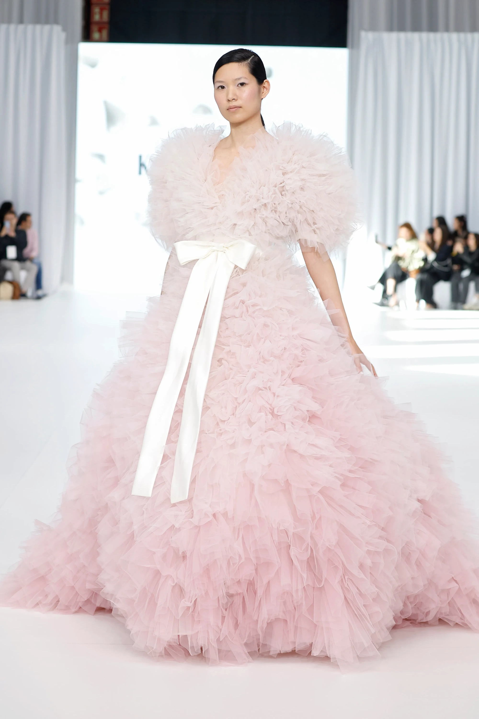 Barcelona Bridal Fashion Week podyumunda, pembe tüllerden kat kat fırfırlarla tasarlanmış, omuzlardan yere kadar uzanan abartılı hacimli bir İlkbahar gelinlik koleksiyonu parçası, belindeki uzun beyaz saten kurdele fiyonk detayıyla romantik ve masalsı bir görünüm sergiliyor.