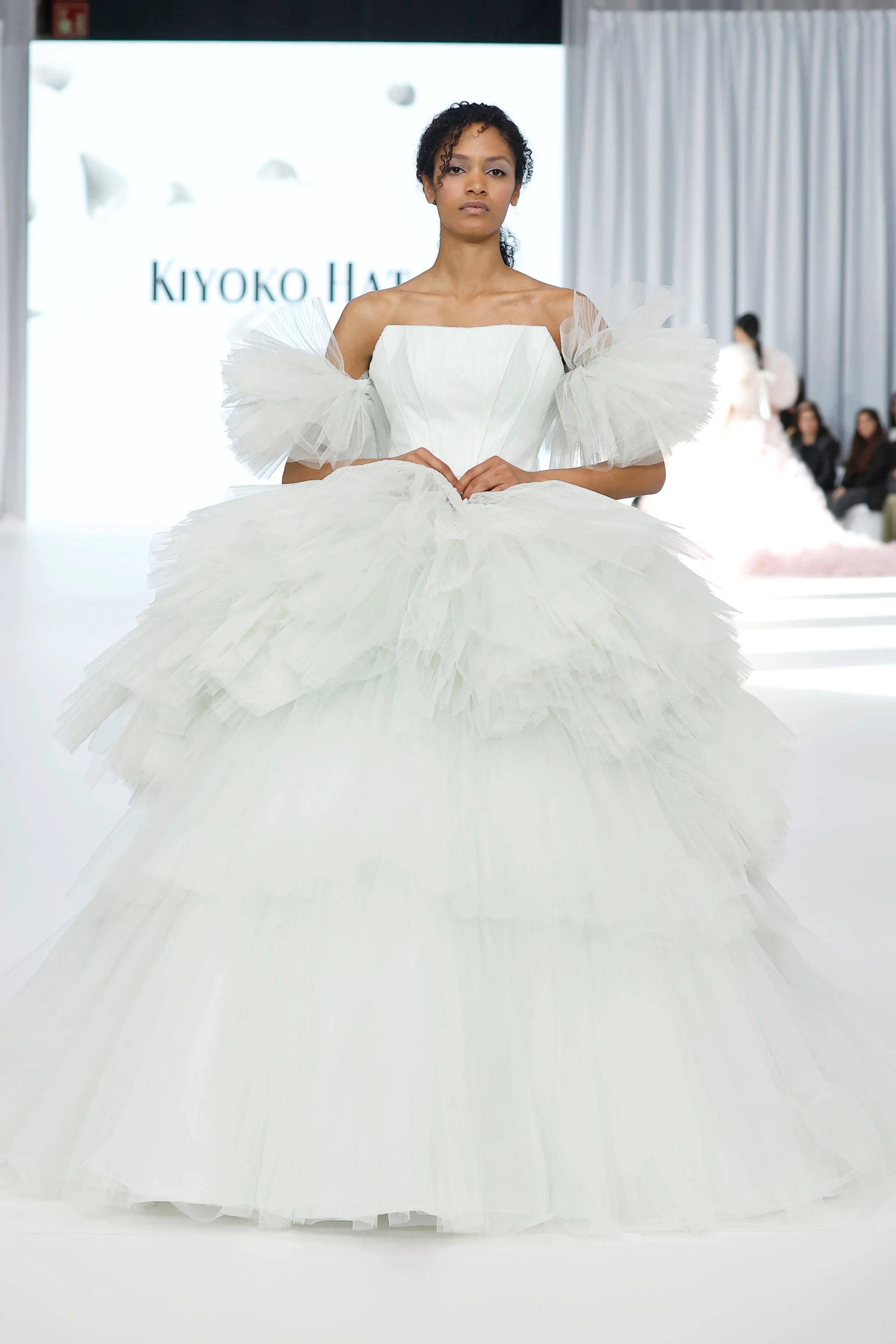 Podyumda duran bir model, straplez korse üst, omuzlardan ayrık, katmanlı kabarık tül kolları ve çok katlı, hacimli beyaz tül eteğiyle dikkat çeken bir gelinlik giyiyor; Kiyoko Hata İlkbahar 2027 gelinlik koleksiyonundan bu gösterişli tasarım, zanaatkârlık ve modern formu bir araya getiriyor.