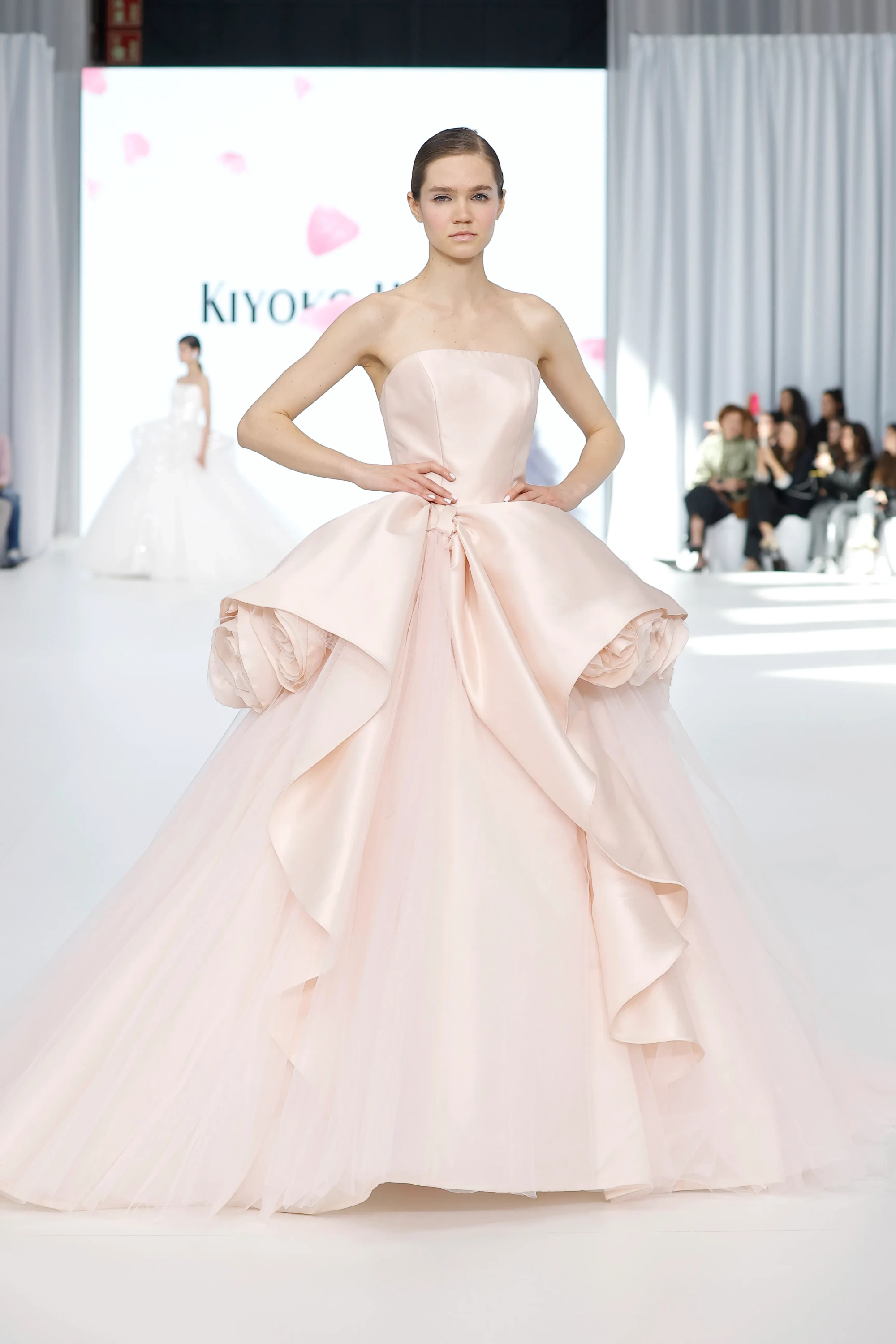 Model, beyaz podyumda elleri belinde poz verirken, Barcelona Bridal Fashion Week'te sunulan bir koleksiyondan, hacimli tül eteği ve büyük, yumuşak çiçek formlarıyla süslenmiş, ustaca drapeli, heykelsi bir beli olan straplez açık pembe bir Kiyoko Hata gelinlik sergiliyor.