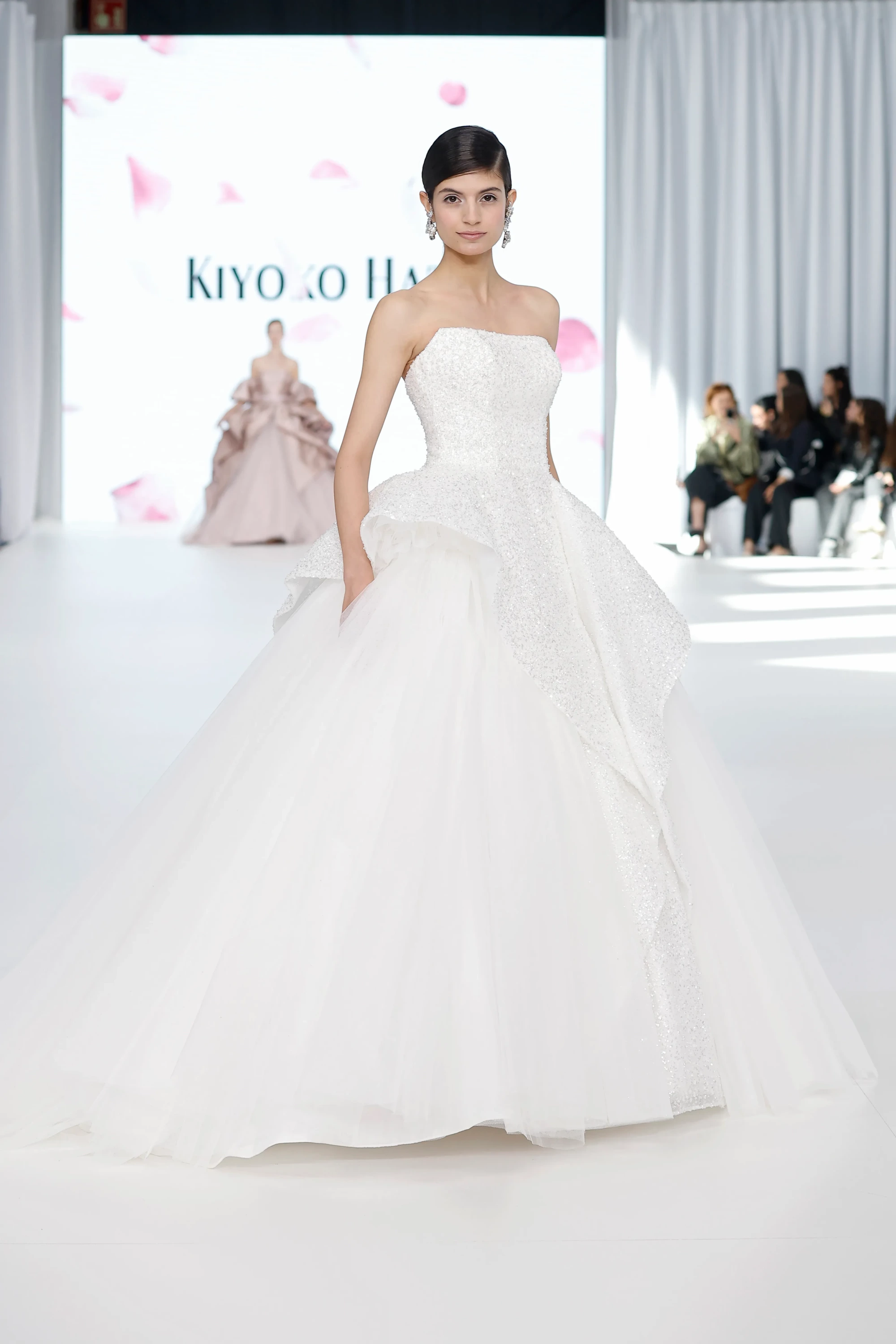 Barcelona Bridal Fashion Week'te sunulan Kiyoko Hata gelinlik koleksiyonundan bir model, straplez, tamamen ışıltılı büstiyerli ve kabarık, katmanlı tül etekli dramatik beyaz bir gelinlikle podyumda tüm zarafetiyle duruyor.