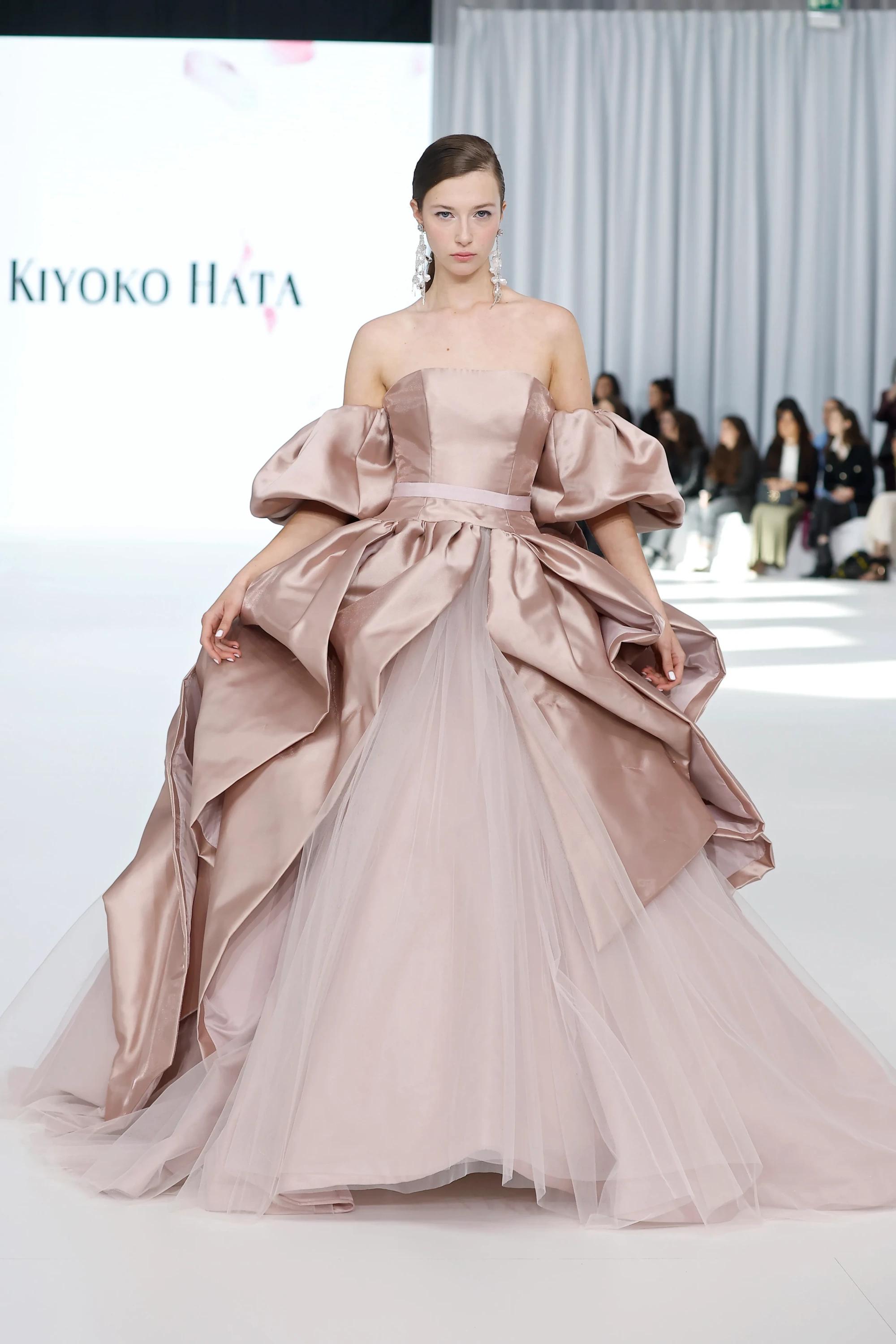 Barcelona Bridal Fashion Week podyumunda, Kiyoko Hata İlkbahar Gelinlik Koleksiyonu'ndan sırtı açık, fırfırlı ve kabarık kolları olan parlak gül kurusu rengi bir gelinlik giyen model, heykelsi üst kısmı ile tül katmanların akışkanlığını birleştiren, zanaatkârlık ve formun dramatik birleşimiyle göz kamaştırıyor.