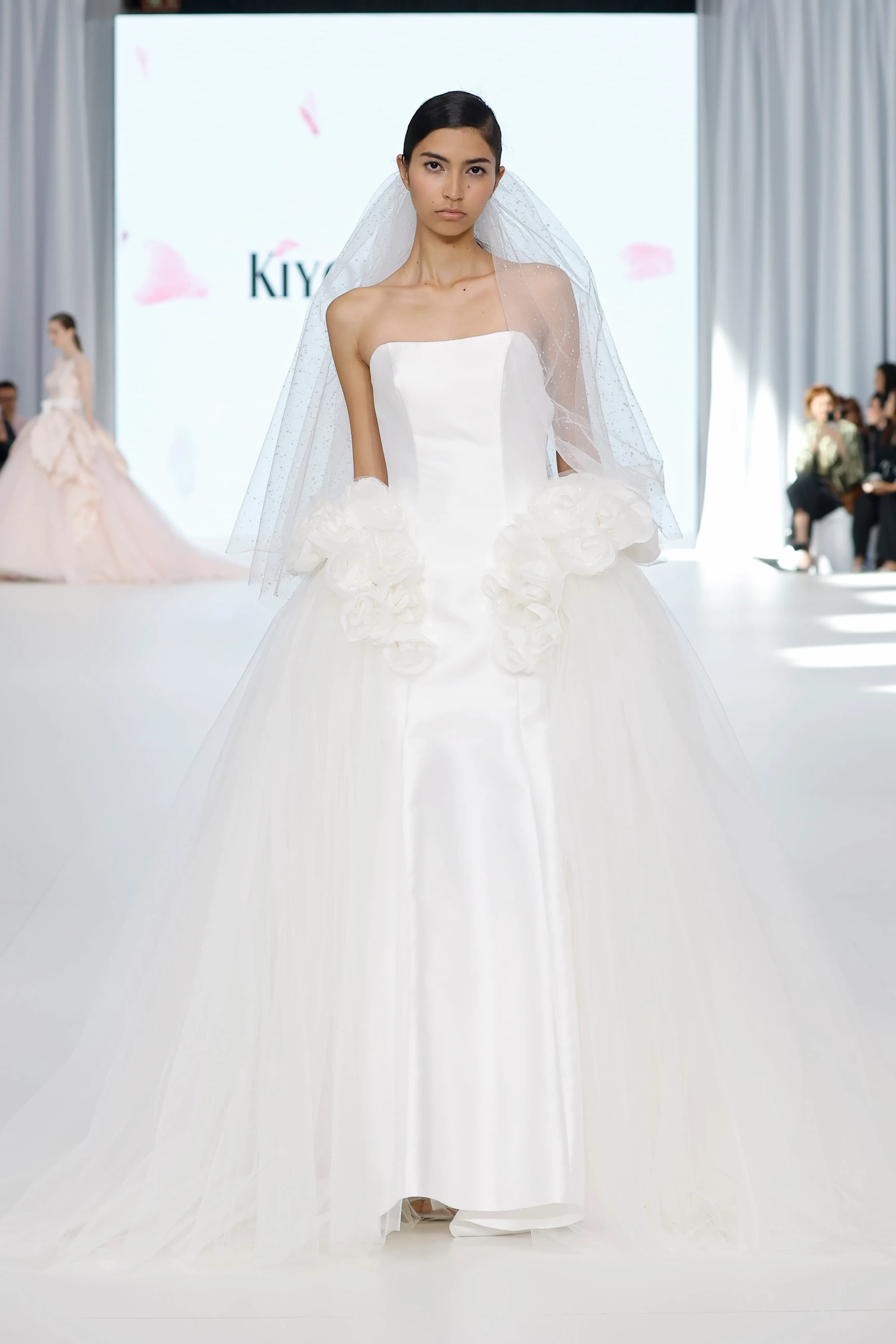 Barcelona Bridal Fashion Week podyumunda, straplez, parlak detaylı tül duvakla tamamlanmış, beli hacimli çiçek formunda aplikelerle hareketlendirilmiş ve etekleri katmanlı tüllerle dramatik bir hacim kazandırılmış beyaz gelinliğiyle bir model.