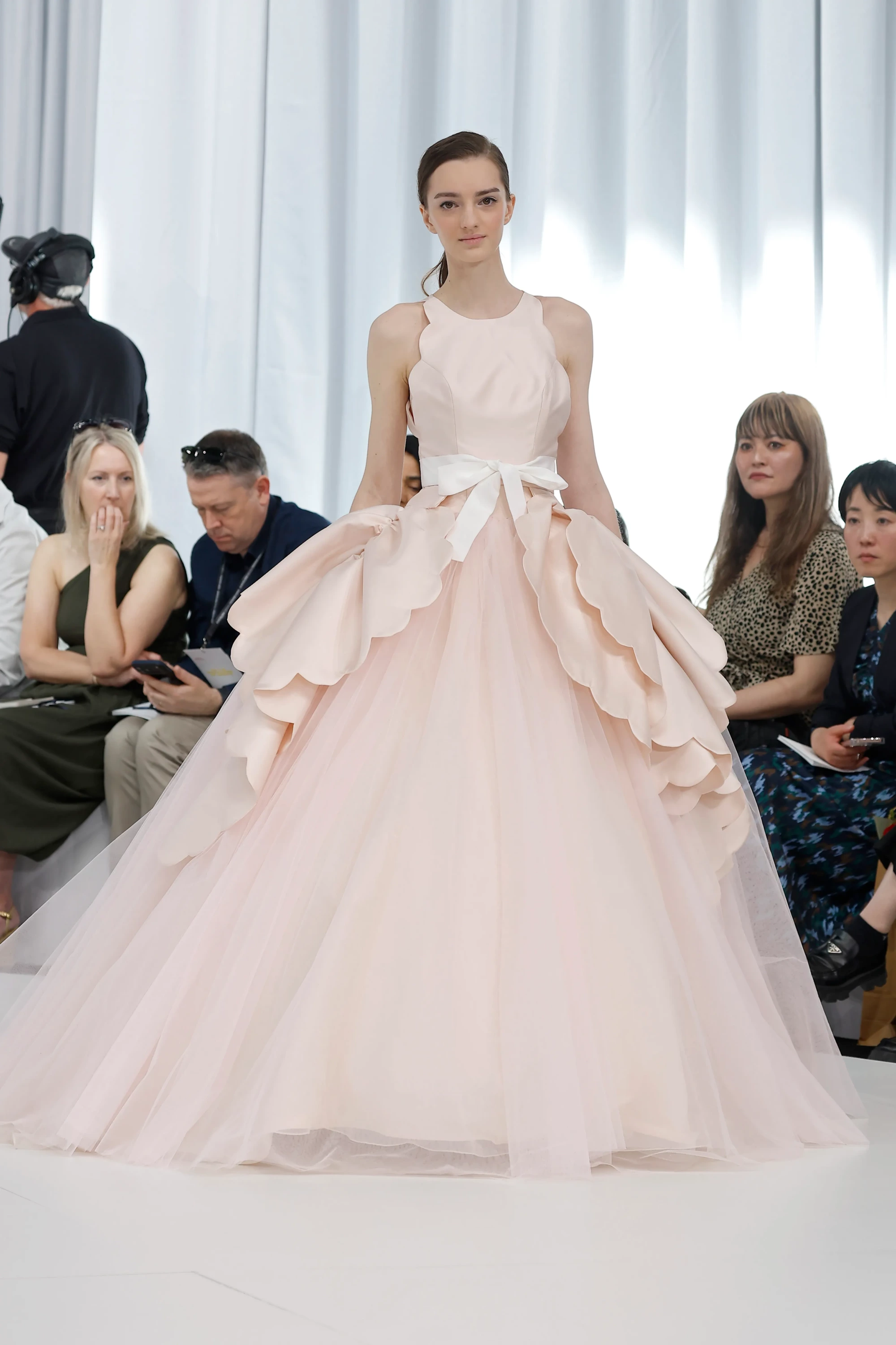Modelin üzerinde, Vaishali Studio İlkbahar 2027 Gelinlik Koleksiyonu'ndan, üst kısmındaki heykelsi dalgalı formları ve belindeki beyaz kurdele detayından sonra transparan, akışkan tül katmanlarla zemine yayılan pastel pembe bir gelinlik belirginleşiyor.