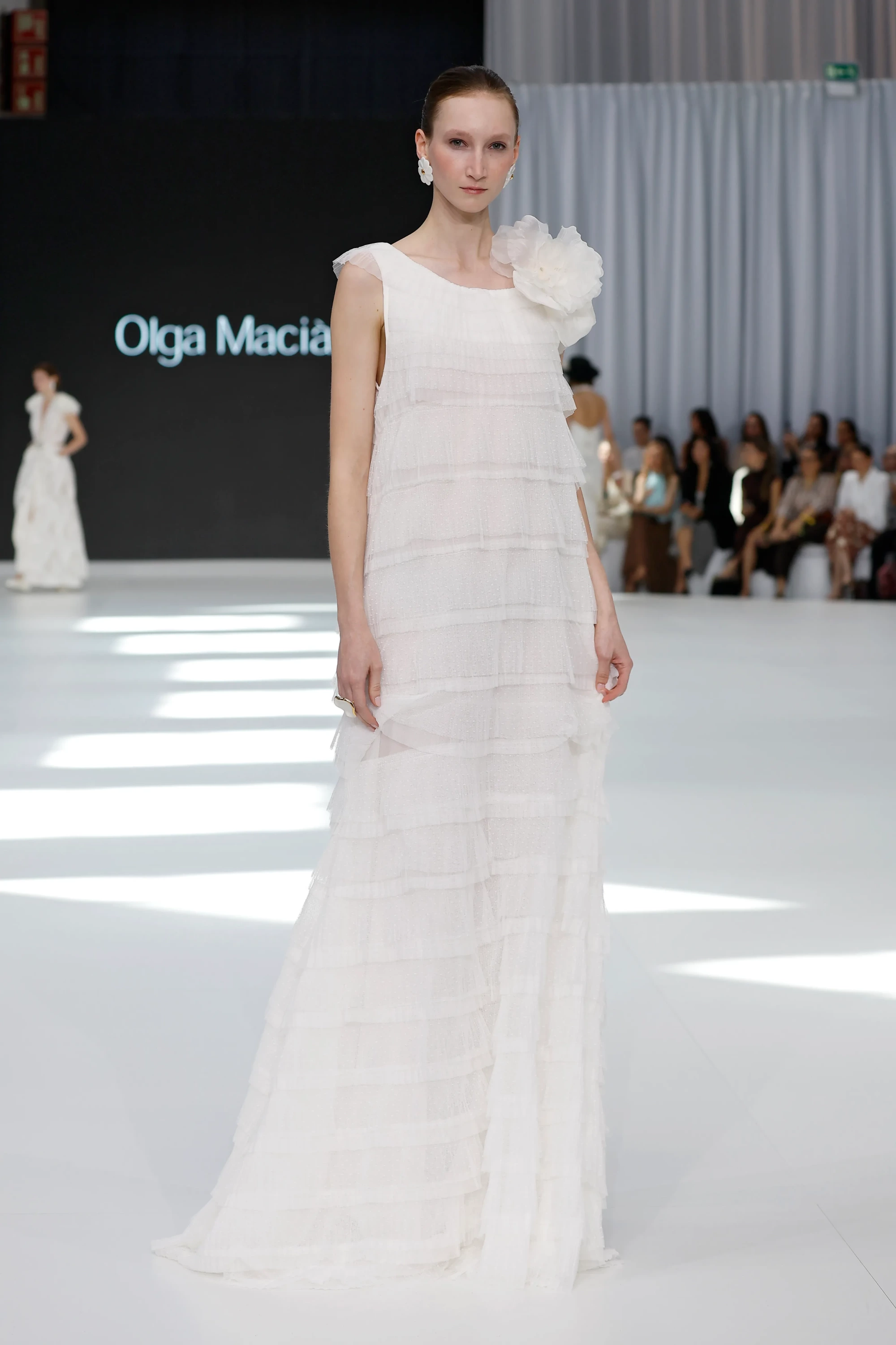 Barcelona Bridal Fashion Week'te Olga Macià İlkbahar 2027 Gelinlik Koleksiyonu'ndan, akışkan kumaşlarla tasarlanmış, katmanlı plumetis fırfırlara ve omzunda büyük çiçek işlemeye sahip uzun beyaz bir gelinlikle podyumda duran zarif bir model.