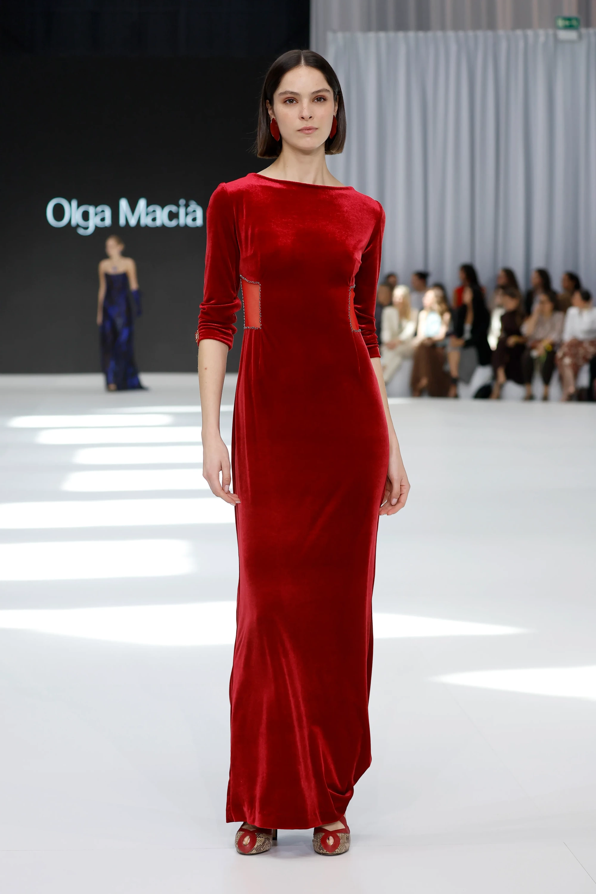 Model, Olga Macià koleksiyonundan, kayık yaka, üç çeyrek kollu, vücudu saran kırmızı kadife uzun bir elbise ile podyumda yürüyor; elbisenin transparan, boncuk işlemeli yan panelleri, İlkbahar 2027 Gelinlik Koleksiyonu'nun modern ve klasik çizgileri harmanlayan flamenko ruhunu yansıtıyor.