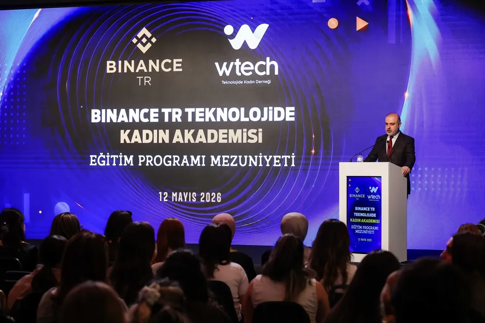 Büyük bir sahnede, arkasında Binance TR ve wtech logosu ile Binance TR Teknolojide Kadın Akademisi Eğitim Programı Mezuniyeti başlığının ve 12 Mayıs 2026 tarihinin yazılı olduğu devasa bir ekranın önünde, takım elbiseli bir adam kürsüde konuşma yaparken, etkinliği dinleyen kalabalık bir katılımcı grubu önden görünüyor.