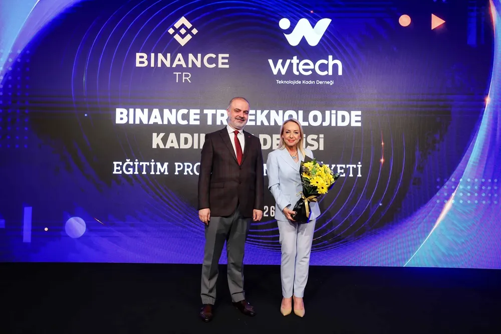 Binance TR ve Teknolojide Kadın Derneği Wtech'in ortaklaşa düzenlediği Eğitim Programı etkinliğinde, arkasında büyük mavi bir ekranda program başlığının yazılı olduğu sahnede, koyu renk takım elbiseli bir beyefendi ve elinde sarı çiçek buketi tutan açık mavi takımlı bir hanımefendi gülümseyerek poz veriyor.