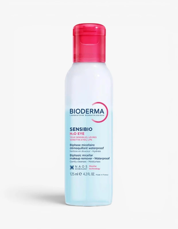 Beyaz bir arka plan üzerinde, pembe kapaklı, Bioderma Sensibio H2O Eye çift fazlı makyaj temizleyici şişesi; hassas göz ve dudaklar için özel olarak formüle edilmiş, suya dayanıklı makyajı nazikçe arındırmayı vadeden, şeffaf üst ve açık mavi alt kısımdan oluşan 125 ml'lik berrak sıvı içeriğini sergilemektedir.