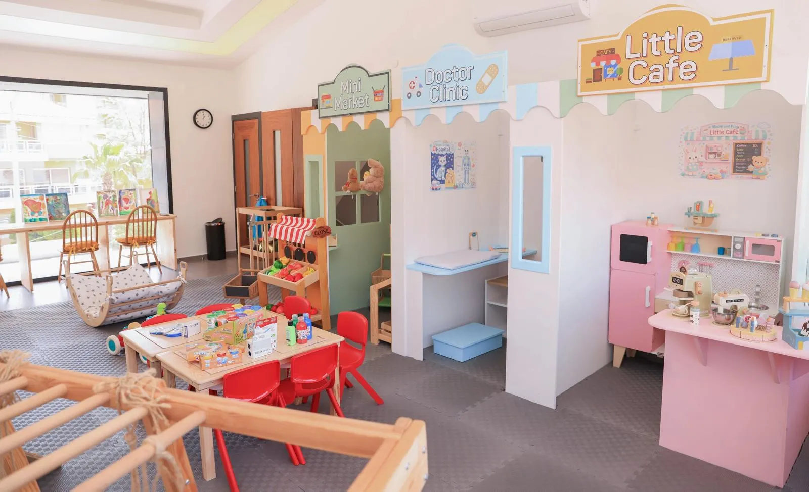 Aydınlık ve ferah bir iç mekanda, çocukların rol yapma becerilerini geliştirebileceği; içinde mini market, doktor kliniği ve pembe renkli küçük kafe gibi çeşitli temalı oyun evleri ile oyuncak yiyeceklerle dolu bir masa bulunan, eğitici ve eğlenceli bir oyun alanı görülüyor.