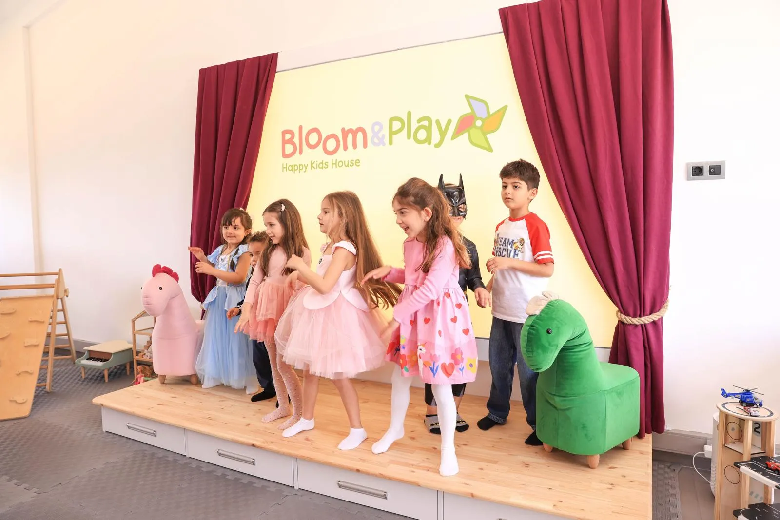 Bloom & Play Happy Kids House sahnesinde prenses, peri ve Batman gibi çeşitli kostümler giymiş neşeli çocuklar enerjik bir şekilde dans edip eğlenirken, arkalarındaki Bloom & Play logosu ve yandaki bordo perdeler bu keyifli etkinliğin canlı atmosferini tamamlıyor.