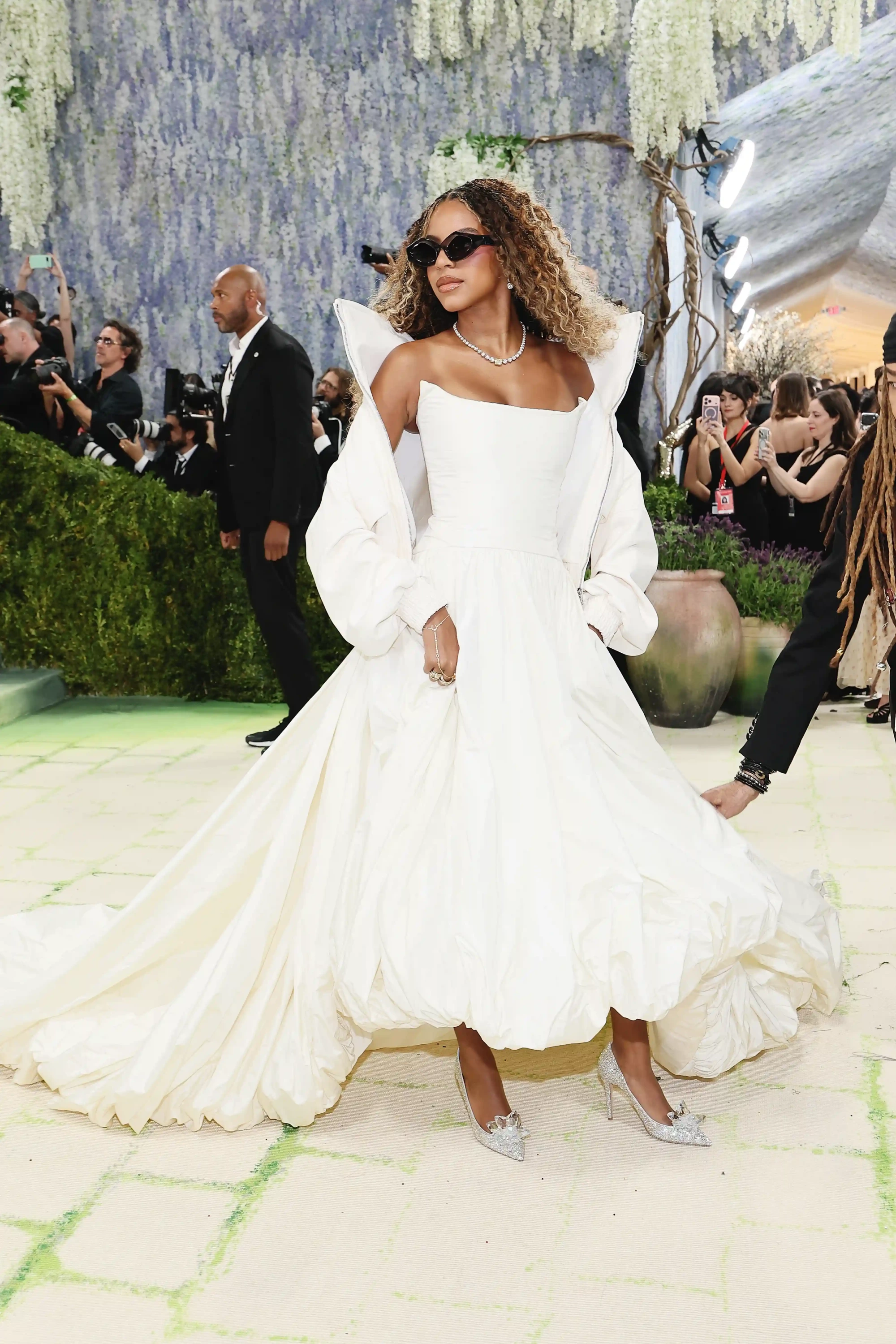 Beyoncé, 2026 Met Gala kırmızı halısında, omuzları açık, kabarık etekli, uzun kuyruklu krem rengi elbisesi ve üzerine düşen hacimli ceketiyle, pırıltılı takıları ve koyu renk güneş gözlükleriyle adeta yaşayan bir moda ve kostüm sanatı eseri gibi büyüleyici bir görünüm sergiliyor.