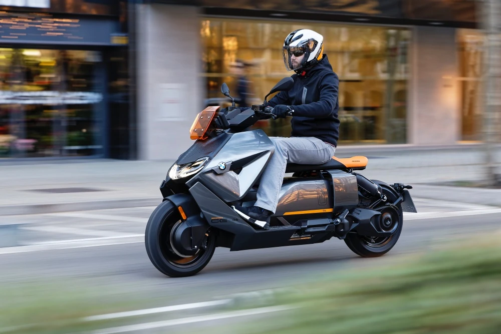 Beyaz kask ve siyah ceket giymiş bir sürücü, gri gövdesinde turuncu vurguları olan fütüristik görünümlü bir BMW CE 04 elektrikli scooter ile şehir içinde hızla ilerliyor ve dinamik bir şehir içi motosiklet sürüş anını gösteriyor.